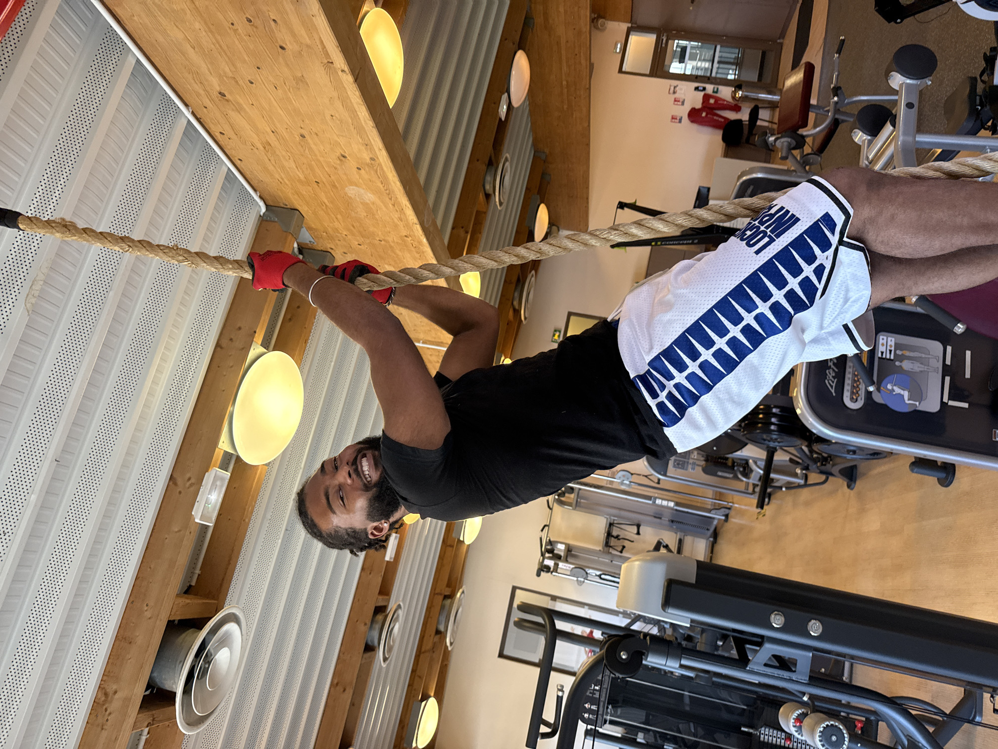 The image shows a man hanging from a rope in a gymnasium. He is wearing red gloves, a black t-shirt, and white shorts with blue trim. The gym has exercise equipment in the background and a wooden ceiling with lights. The location is identified as Sarcelles, France.