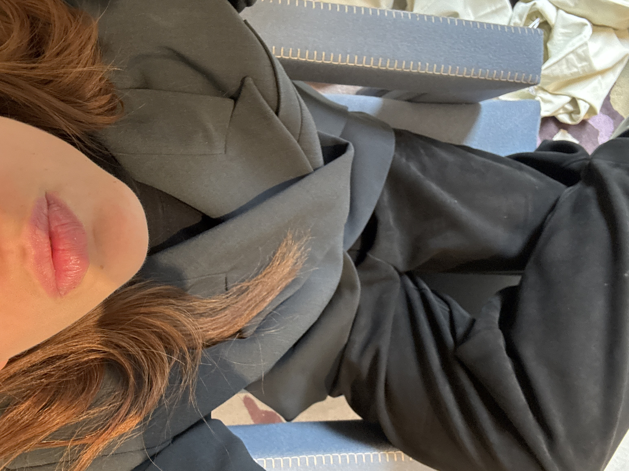 A close-up, slightly angled overhead shot captures part of a person with red hair and visible lips. They are wearing a dark gray or black jacket, possibly a blazer. To the right, there are what appear to be black pants or draped material. A light blue box with light-colored stitching detail is partially visible. In the background, there are hints of crumpled light-colored fabric. There are no indications of Paris, France in the visible elements.