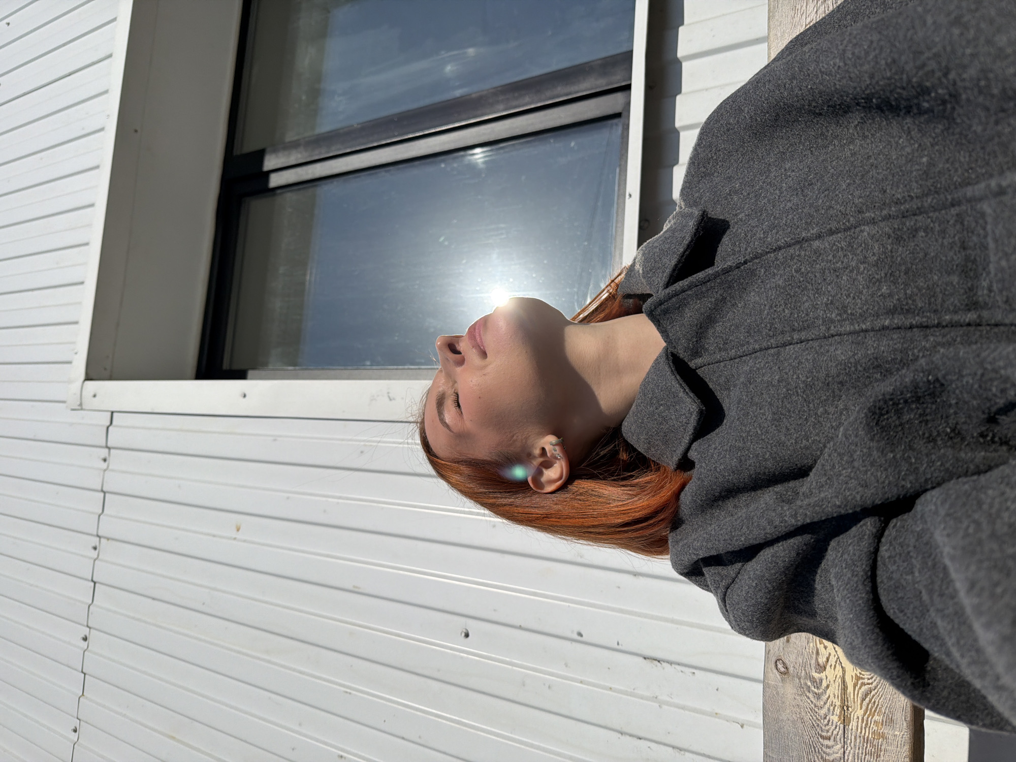 A woman with red hair, wearing a gray coat, is leaning back against a white, corrugated metal building with a window. The sun reflects off the window directly onto her face. The location is Chaikovsky, Russia.