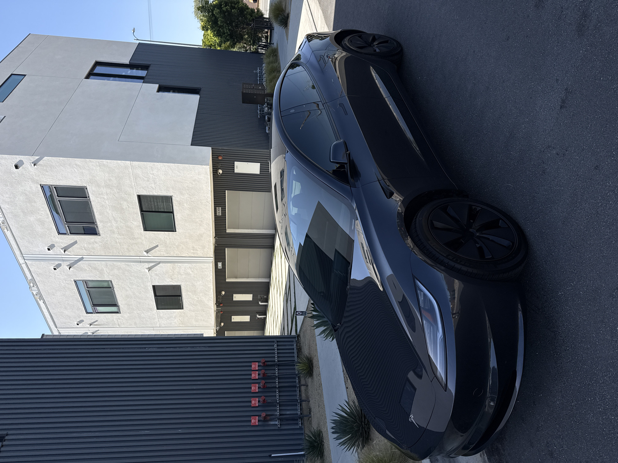This image, captured in Los Angeles, United States of America, presents a dark grey Tesla Model S parked on an asphalt street alongside a modern, multi-story building. The bright, clear blue sky suggests a sunny day, likely midday or early afternoon, with distinct shadows cast on the scene.
The Tesla, positioned prominently on the right side, faces slightly away from the viewer, showcasing its sleek design and dark wheels. Reflections of the surrounding architecture and sky are visible on its body.
The building to the left exhibits contemporary architectural styles, featuring a combination of light grey stucco and dark grey corrugated metal paneling. It has multiple levels with various rectangular windows. The number "11300" is clearly visible on a dark section of the building, serving as the address marker. At ground level, next to what appears to be garage doors, there are several red-capped connections for a fire suppression system, each bearing a "DANGER" label.
A narrow landscaped strip runs between the car and the building, featuring several small, spiky agave or succulent plants set in gravel. A small white "NO PARKING" sign is faintly visible among these plants. No people or activities are depicted, giving the scene a quiet and still atmosphere.