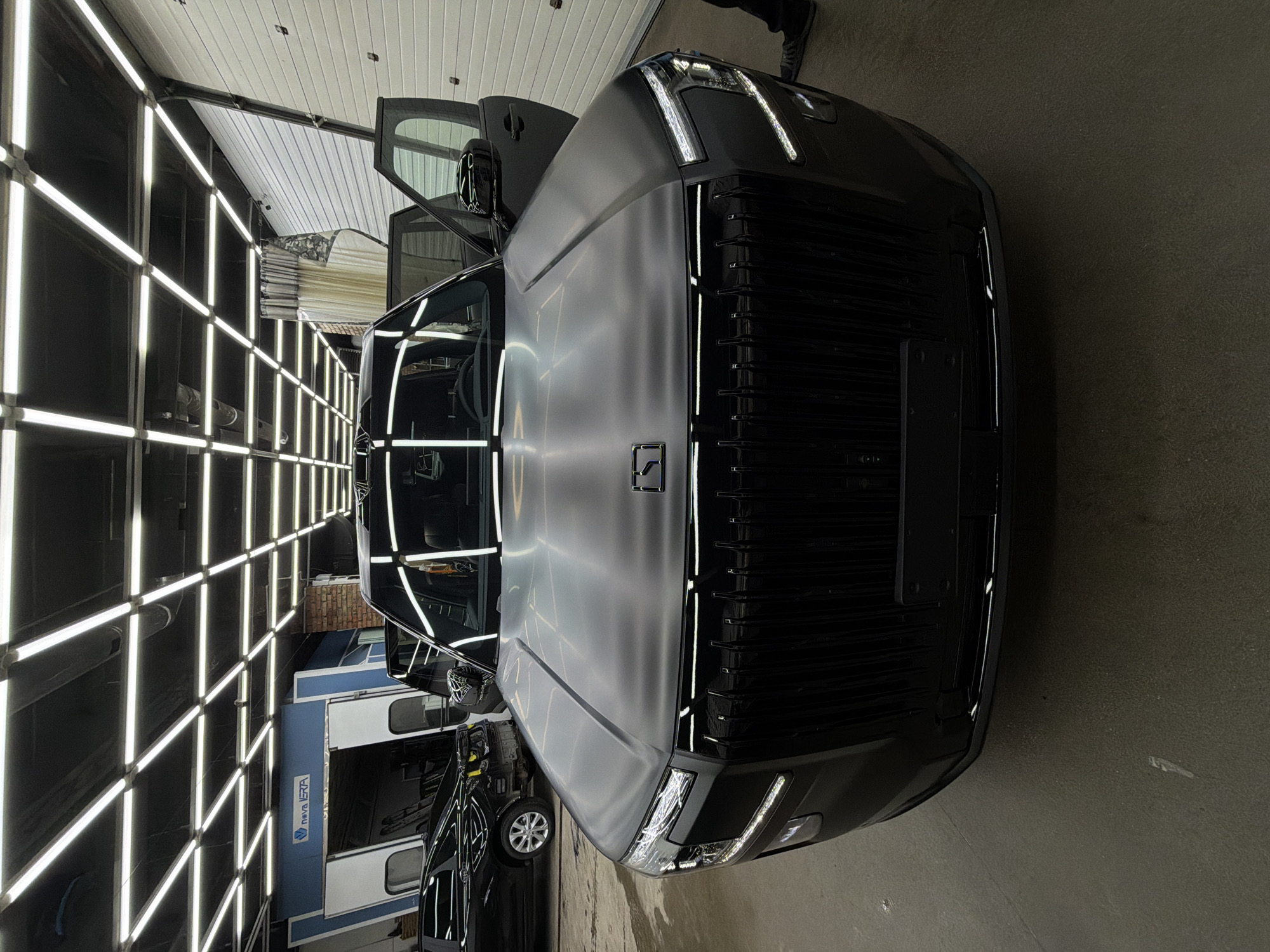 A black vehicle is parked inside a building with bright, squared lighting on the walls and ceiling, giving a grid-like effect. The vehicle appears to be a new or recently detailed car, with a large front grill and open driver's side door. Another car is partially visible behind the primary vehicle. A person's legs are visible in the upper right corner of the image. Signage for "Nova Era" is on the wall in the background. While there is no definitive evidence to determine the exact location in Krasnoyarsk, Russia, it can be reasonably assumed the image was taken in an auto detailing shop, car showroom, or vehicle service center.
