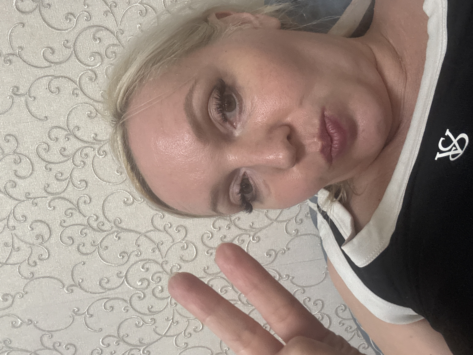A woman with blonde hair and makeup is posing for a photo, holding up two fingers in a peace sign. She is wearing a black and white top. The background appears to be a wall with a decorative pattern. The image was taken in Komsomolsk-on-Amur, Russia.