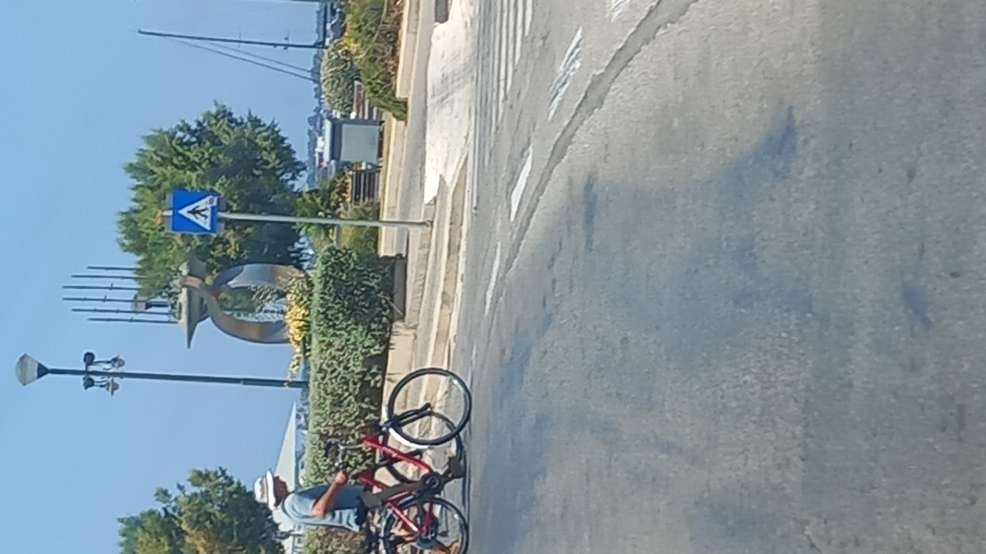 A man wearing a white hat and a light blue t-shirt is standing next to a red bicycle, holding its handlebars. The bicycle's front wheel is turned towards the left. In the background, there is a large, abstract metal sculpture with curved elements and vertical poles. Adjacent to the sculpture, a blue pedestrian crossing sign with a white pedestrian symbol is visible on a pole. The scene is set against a clear blue sky and a backdrop of green trees and bushes. The ground is paved with asphalt, and a pedestrian crossing with white stripes is marked on the road in the foreground. The location appears to be near a waterfront, with a sailboat mast visible in the far distance. This image was taken in Kos, Greece, specifically at the harbor area near the Eleftherias Square.