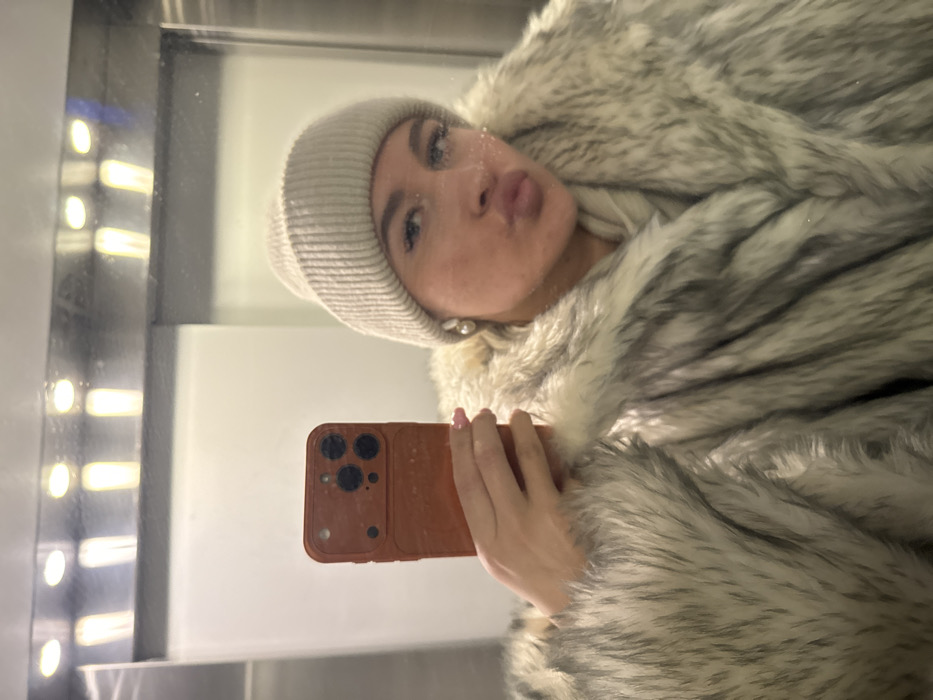 The image displays a reflection of a female individual captured in a mirror selfie, situated within an indoor environment characterized by highly reflective metallic or mirrored surfaces. The specific location within Odintsovo, Russia, cannot be determined from the visual evidence.
The subject is centrally framed, wearing a light beige, ribbed knit beanie and a voluminous faux fur coat with a distinct pattern of mixed grey and white fibers. A light-colored garment, possibly a scarf or turtleneck, is visible around her neck beneath the coat. Her face shows pouted lips; small, reflective glitter-like particles are present on her upper lip and around her nose. She wears light-colored, spherical stud earrings.
In her right hand, the subject holds a smartphone, encased in a dark orange, textured protective cover. The rear of the smartphone displays a prominent triple-lens camera array and an additional sensor. Her visible fingernails are painted a light pink hue.
The background, visible as a reflection, indicates an enclosed space. Above the subject, a row of four oblong light fixtures emitting a warm, yellowish light is reflected. Vertical lines and a lighter, possibly off-white, panel are also discernible in the reflective surface, consistent with an elevator interior or a mirrored hallway.