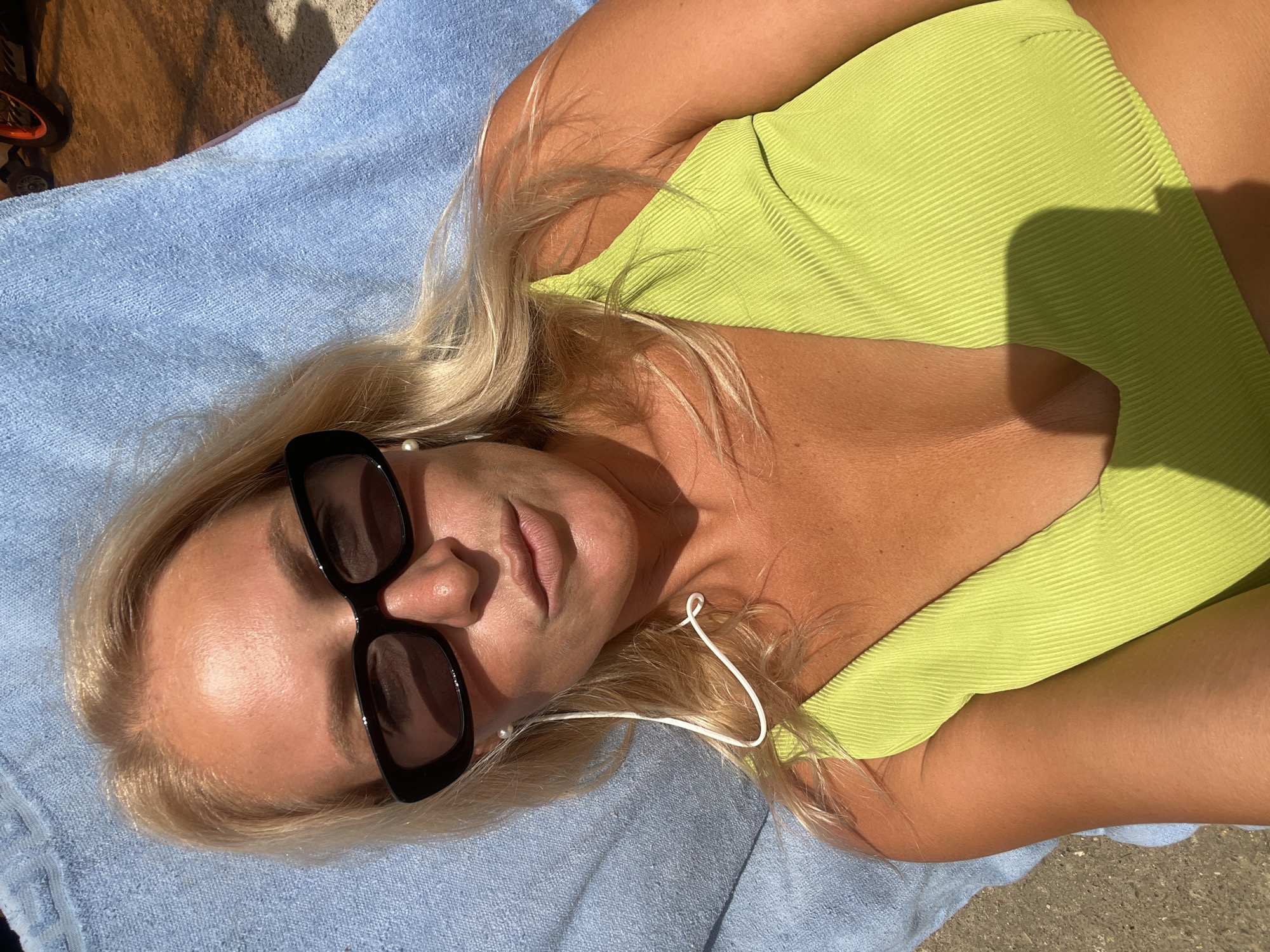 A woman with blonde hair and sunglasses lies on a blue towel. She is wearing a green bikini top. Her eyes are looking at the camera, and her lips are slightly parted. She has white earbuds in her ears. The environment is outdoors, with sunlight casting shadows. It is not possible to determine the location in Baranovka, Russia from this image.