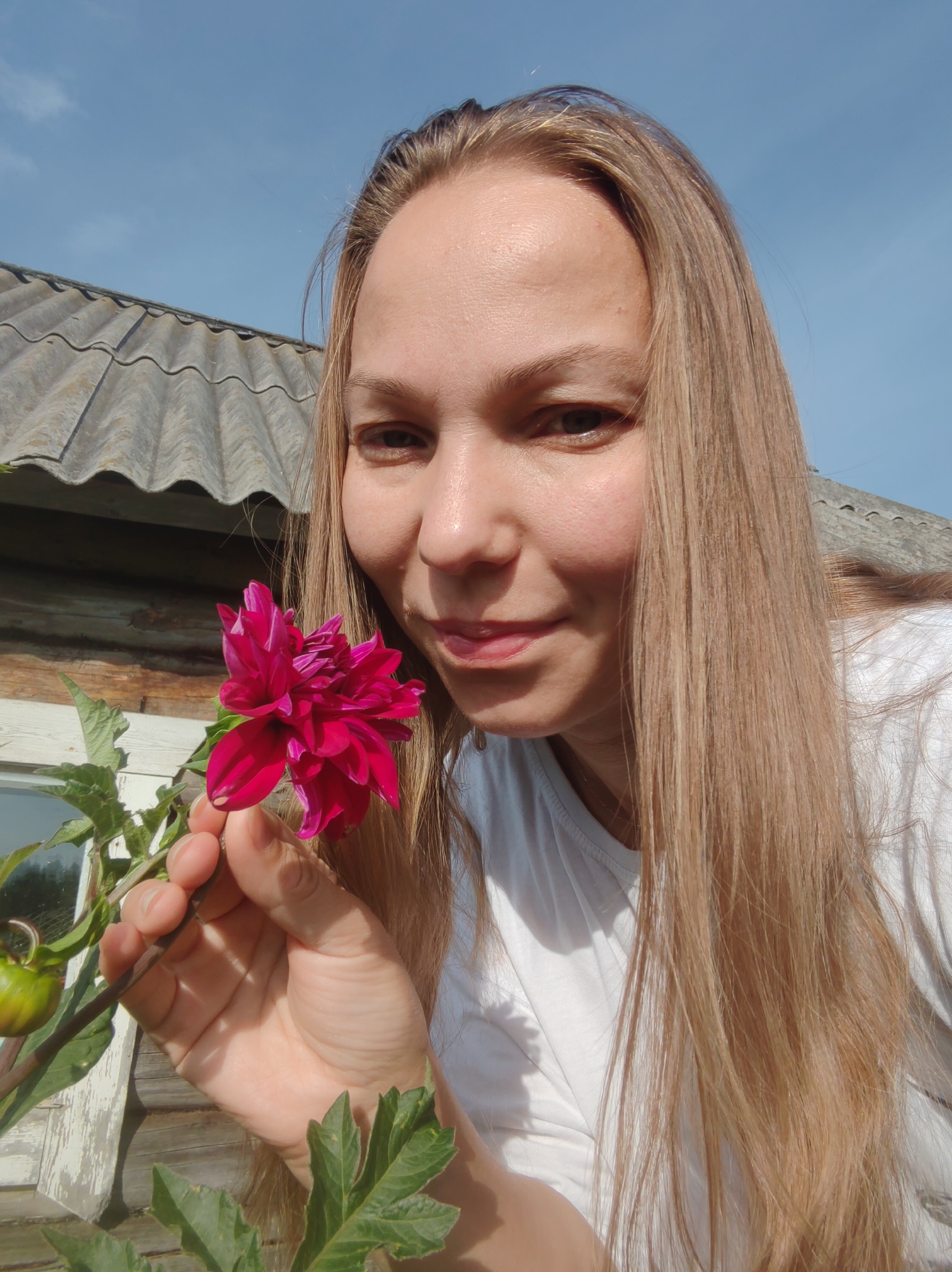 The image shows a young woman with long, blonde hair holding a bright pink flower. She is wearing a white t-shirt. In the background, there is a wooden house with a grey, corrugated roof. The setting appears to be outdoors, possibly in a rural area. The sky is blue with a few wisps of clouds. The woman's left hand is holding the stem of the flower, and her face is close to it, as if she is smelling it or showing it to the camera. Her expression is soft and smiling. A green bud is visible on the plant to the left of the flower. The lighting suggests it is daytime, with sunlight illuminating the scene. The house's wooden planks are visible, some weathered and showing their grain. The roof has a textured, wavy appearance. The overall impression is one of a casual, sunny day in a natural setting. The location of Чухлэм, Russia, is mentioned as part of the context.