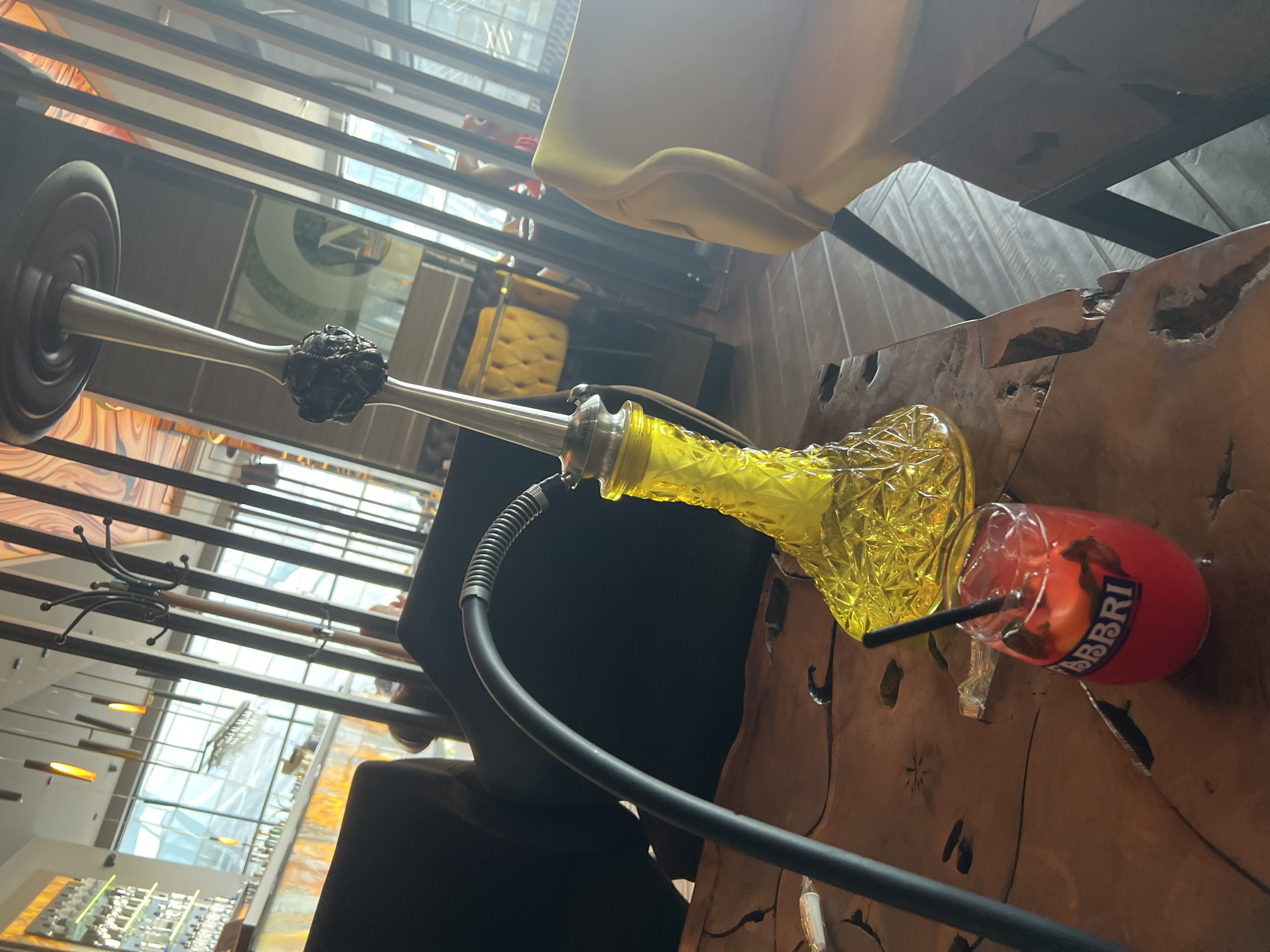 An overhead, tilted shot captures a leisure scene featuring a hookah and a cocktail on a rustic wooden table within a modern indoor setting, likely in Burgas, Bulgaria.
The central objects are a bright yellow, faceted glass hookah base with a metallic stem and a black ribbed hose, and a vibrant red cocktail. The cocktail, served in a stemmed glass, contains ice, what appears to be mint leaves or other herbs, an orange slice, and a black straw. The brand "FABBRI" is clearly visible in white letters on the side of the cocktail glass. A partial word, possibly "MARKLAUD" or "MARKLAND," is visible on the metal stem of the hookah.
The table is crafted from dark, irregular wood, featuring natural edges and carved-out sections, giving it a raw, organic aesthetic. In the background, reflections from large windows or glass partitions suggest an expansive space, possibly with an outdoor view or another part of the interior. Horizontal architectural slats or beams are prominent in the upper and middle portions of the image. Yellow upholstered chairs are also visible, contributing to the lounge-like ambiance. Further in the background, suspended lighting fixtures can be seen, along with a black metal coat rack.
The overall impression is one of relaxation and indulgence in a stylish, well-lit environment, suggesting an enjoyable social gathering or individual leisure time during what appears to be daytime, given the natural light filtering through the windows.