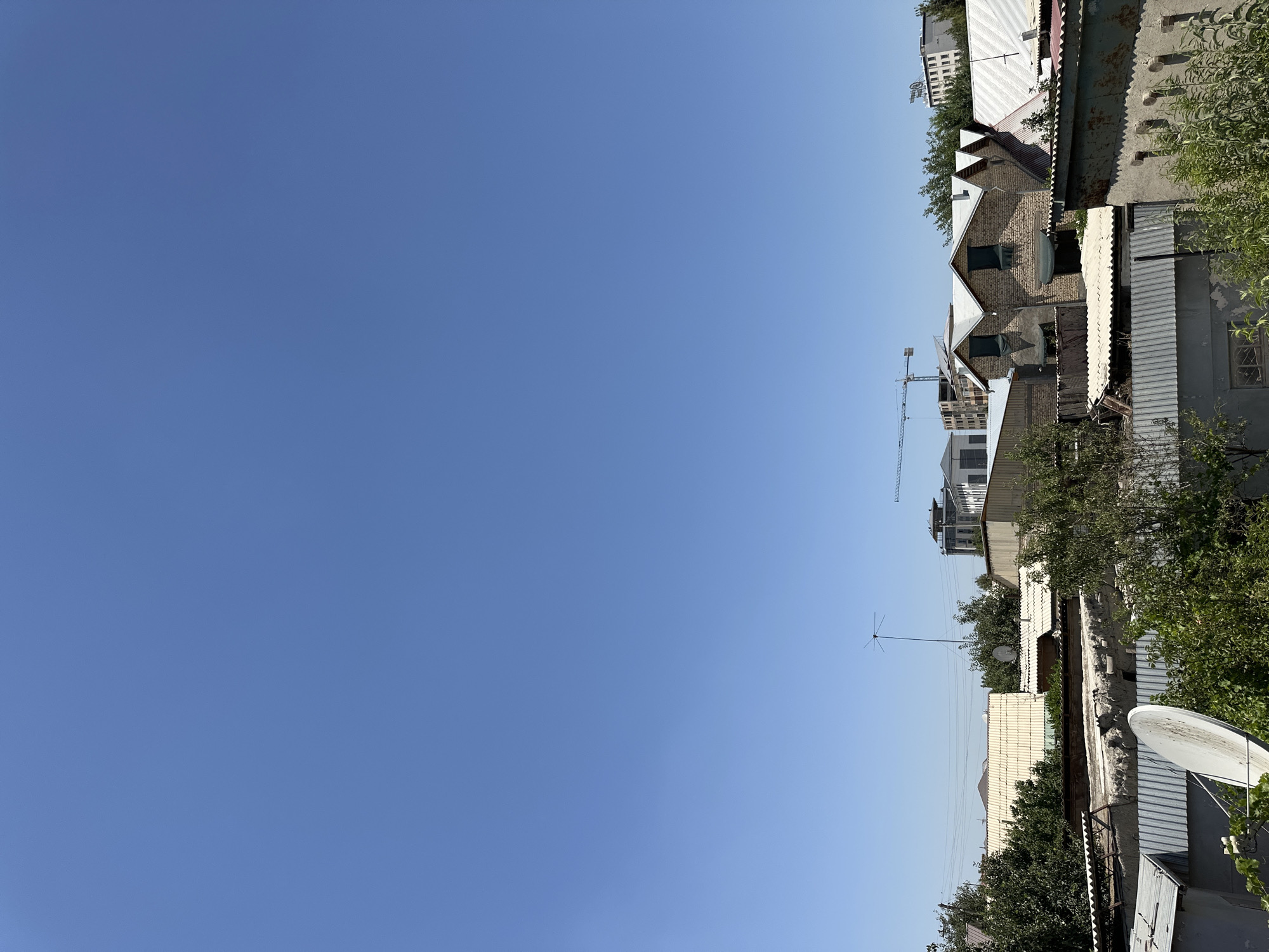 The image depicts a clear, bright blue sky over a cityscape. The foreground is dominated by a collection of low-rise buildings with corrugated metal and tiled roofs, interspersed with lush green trees and foliage. In the mid-ground, more substantial brick and concrete structures are visible, some appearing to be under construction with cranes in the background. A prominent satellite dish is situated in the lower left portion of the frame. The overall scene suggests an urban environment in Kogon City, Uzbekistan, characterized by a mix of residential and developing construction.