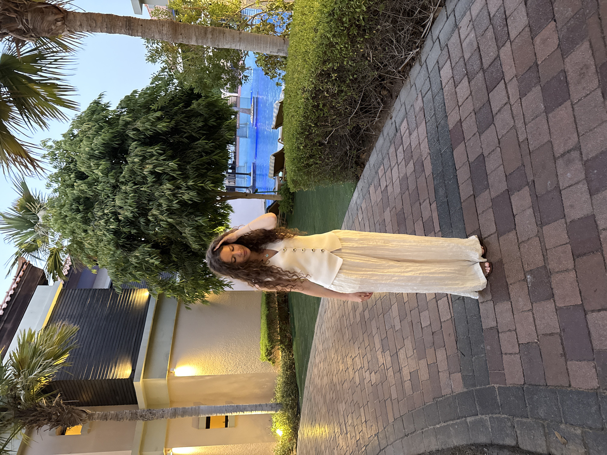 A woman with long curly brown hair stands on a brick pathway in front of a resort in Dubai, United Arab Emirates. She is wearing a cream-colored vest with gold buttons and matching cream-colored wide-leg pants. Her left hand is touching her head, and her eyes are downcast. She is barefoot.
The resort features a building with a cream-colored exterior and dark wood paneling, illuminated by warm-toned lights. Palm trees and other lush greenery surround the area. In the background, a bright blue swimming pool is visible with lounge chairs nearby. The sky is a pale blue, suggesting it is either early morning or late evening. The pathway is made of red and gray bricks.