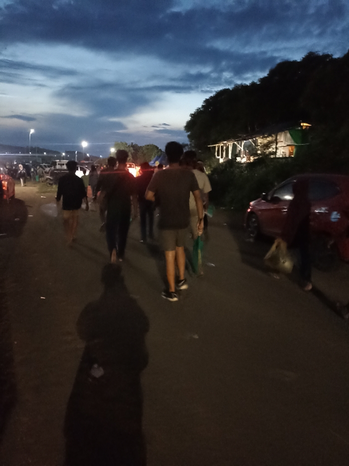 The image captures a scene during dusk or early night, showing a group of people walking along a road in what appears to be a rural or semi-urban area, possibly in Kecamatan Pujut, Indonesia. The sky above is a dramatic deep blue, streaked with lighter clouds, indicating the transition from day to night.
In the foreground, a prominent shadow of the photographer stretches across the asphalt road. Ahead, several individuals are walking away from the camera, mostly seen from behind. Their figures are silhouetted against the ambient light and distant artificial lights. To the left, more people and some vehicles are visible further down the road, with bright lights in the distance suggesting streetlights or other sources of illumination.
On the right side of the road, a red car is parked. Beyond it, partially obscured by dark trees, is a rustic structure with a slanted roof, illuminated from within, suggesting a small dwelling or makeshift shelter. The overall impression is one of a community or group moving along a pathway as evening descends. No visible text is present in the image.