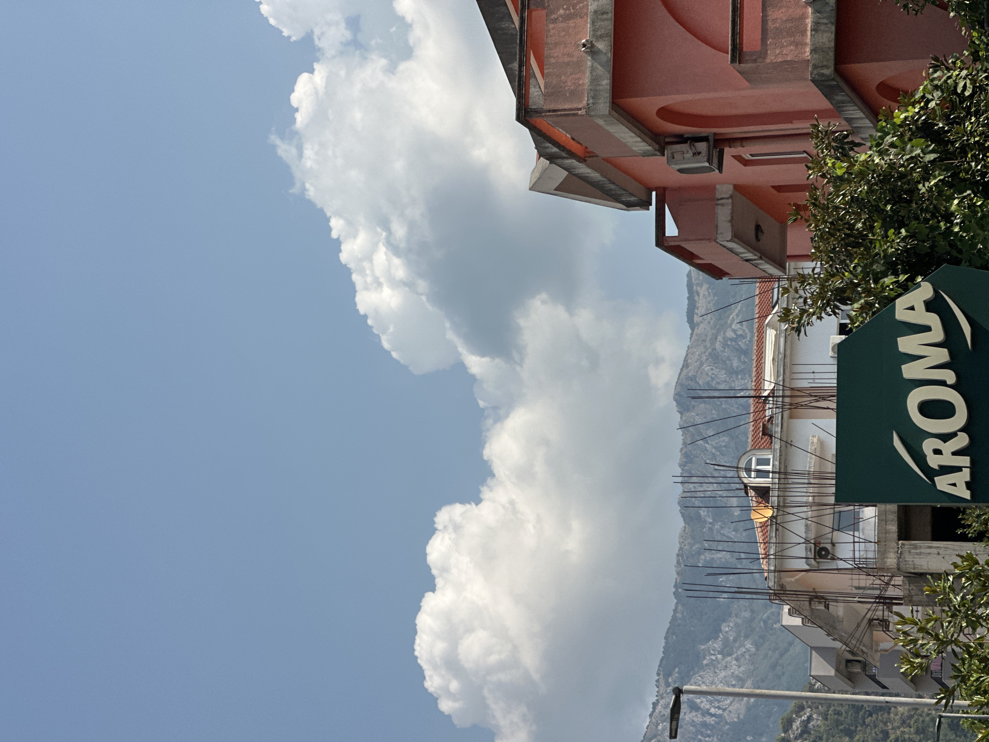 The image shows a street scene in Budva, Montenegro. In the foreground, a green sign with the white logo "AROMA" is visible, partially obscured by lush green foliage. Behind the sign, several buildings are visible. To the right, a prominent pink building with arched windows and balconies stands out. To the left of the pink building, another building is under construction, with exposed rebar and a white facade. Further in the background, a street lamp is positioned to the left of the frame. Majestic mountains form the backdrop to the scene, with a clear blue sky and fluffy white clouds overhead. The lighting suggests it is daytime.