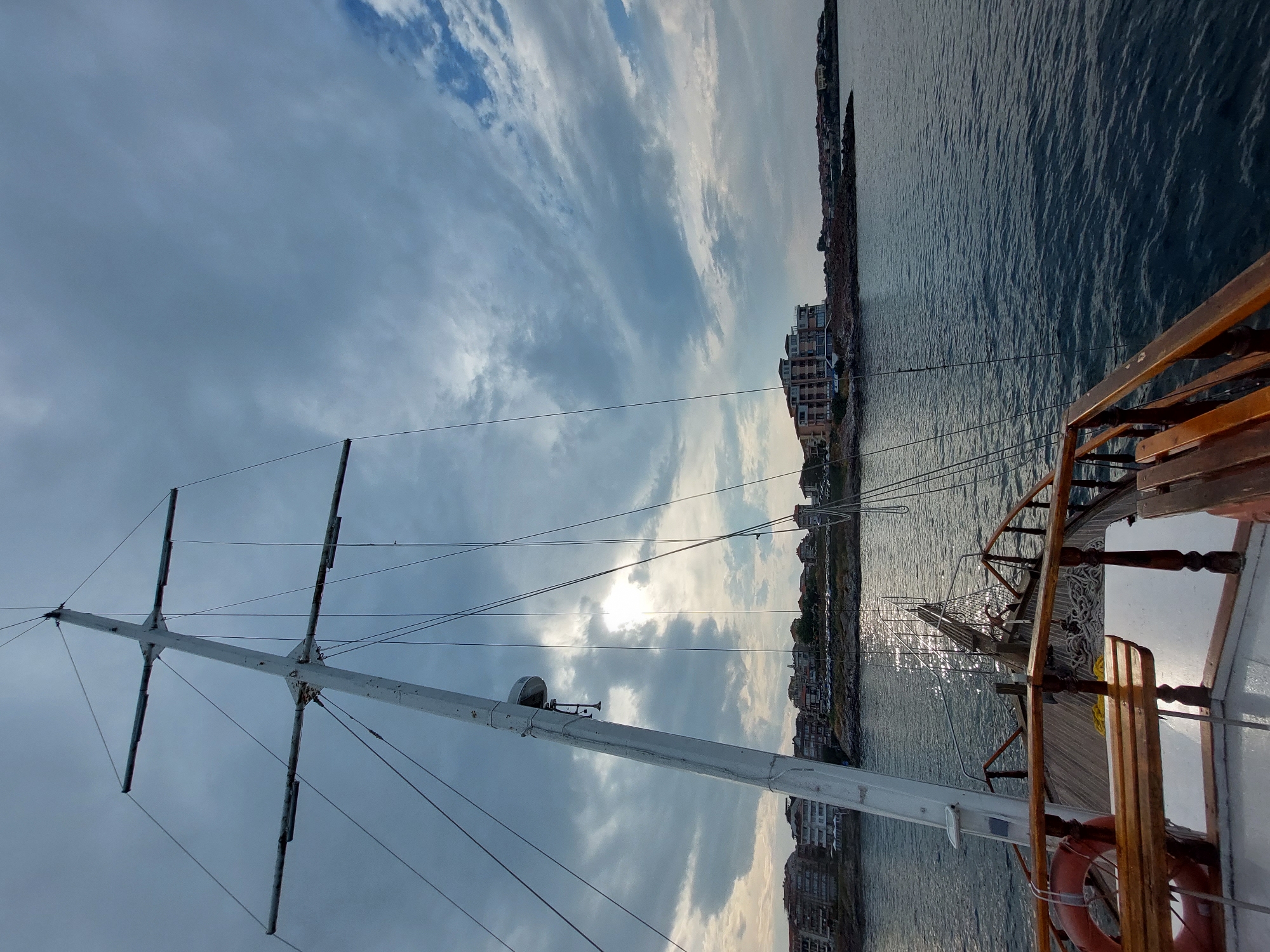 The image captures a view from the deck of a wooden boat sailing on the sea. In the foreground, part of the boat's railing and wooden seating is visible. A white mast with multiple crossbeams and rigging extends upwards into a cloudy sky. The sun is partially obscured by clouds, casting a shimmering reflection on the water. In the background, the coastline of Sozopol, Bulgaria, is lined with buildings, including multi-story structures and smaller houses, situated on rocky terrain. The sea is calm with gentle ripples. The overall scene depicts a nautical perspective of the coastal town under an overcast sky.