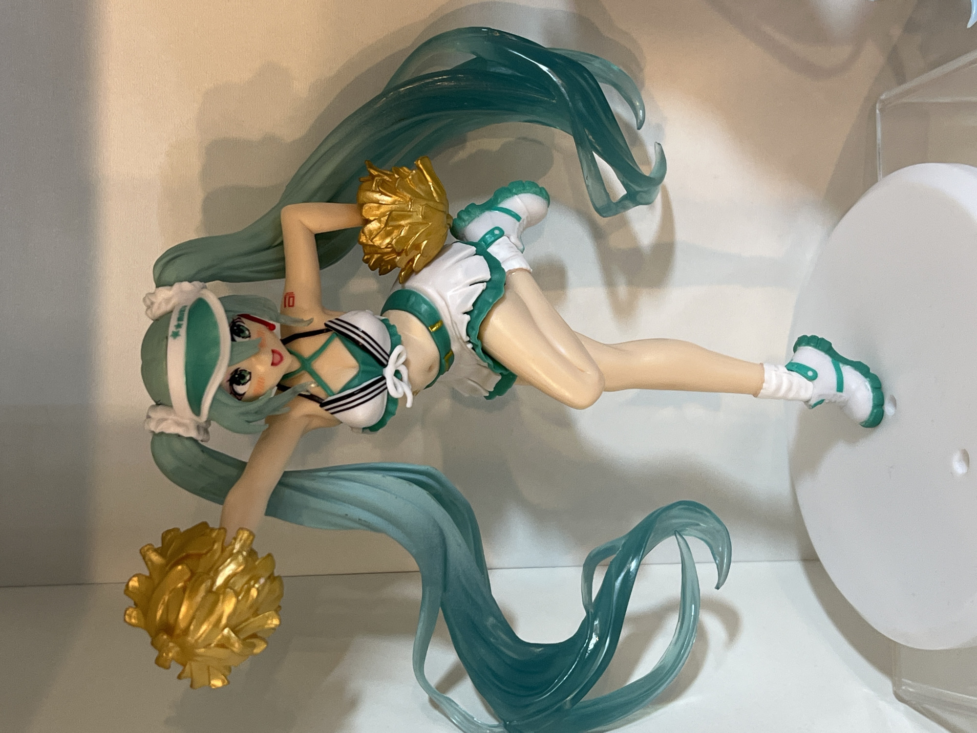 The image shows a full-body, front-facing shot of a Vocaloid Hatsune Miku anime figurine dressed in a green and white cheerleader outfit, complete with a visor and gold pom-poms. The figurine is posed dynamically with one leg bent and the other extended, and its signature teal twin-tails are flowing outwards. The figurine stands on a white circular base, which is placed on a clear display case, and the background consists of a plain, light-colored wall. The lighting is even, casting subtle shadows. The location of the figurine within the city of Arica, Chile, is not determinable from the visual information provided in the image.