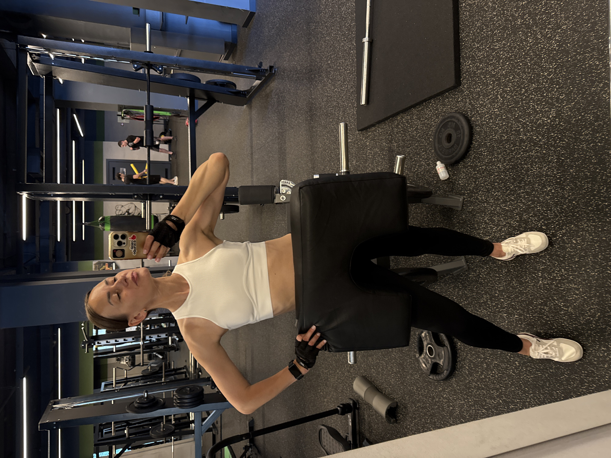 A woman in a white sports bra and black leggings takes a selfie in a gym. She is holding a phone in her right hand and has a black weightlifting belt around her waist, resting on her thighs. She is wearing black fingerless gloves and white sneakers. In the background, there are various gym equipment, including weight racks, barbells, and weights. Two men are visible in the background, one holding a yellow band and the other appearing to be exercising. The gym has a dark floor and a dark ceiling with fluorescent lighting. The overall environment appears to be a commercial fitness facility.