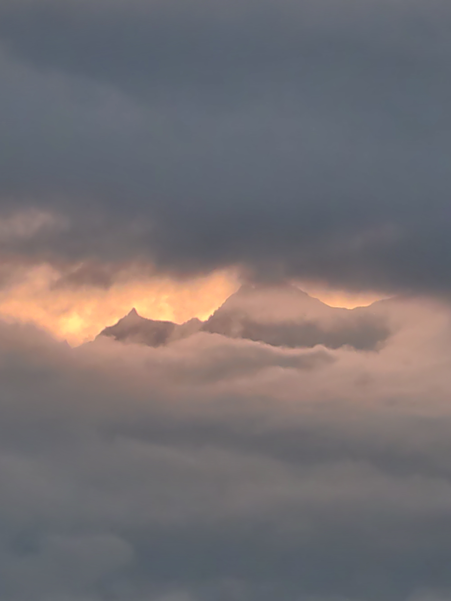 A photograph captures a dramatic sky over the city of Aleo, India. The scene is dominated by dark, moody clouds that obscure much of the sky, creating a sense of an approaching storm or the fading light of dusk. Peeking through a break in the clouds, the rugged peaks of mountains are illuminated by a vibrant, fiery orange glow, suggesting the setting sun or a powerful light source behind them. This glowing aperture in the clouds creates a striking contrast with the somber tones of the surrounding cloud cover. The mountains themselves appear silhouetted against this intense light, their jagged edges sharply defined. Wisps of lighter, pink-tinged clouds drift in front of and around the mountains, softening their appearance and adding depth to the scene. The overall impression is one of natural grandeur and the awe-inspiring power of the elements.