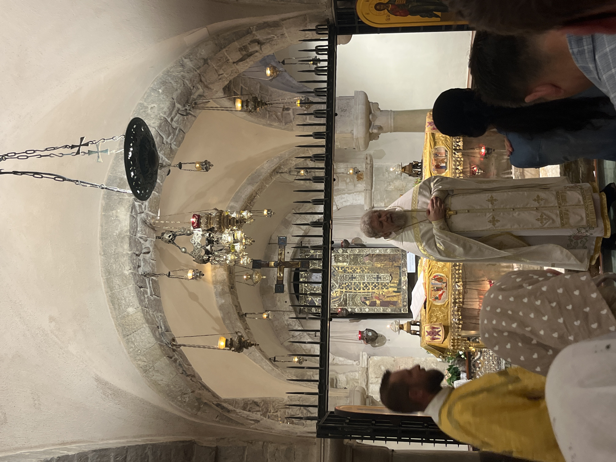 The image depicts an interior scene, likely a church or religious sanctuary, with an arched stone ceiling and multiple hanging lamps. In the foreground, on the right, a priest in white and gold vestments stands with his hands clasped. To his left, a man in a yellow robe stands facing away from the camera. Several people are visible in the midground and foreground, some facing the priest, others with their backs to the camera. Behind the priest, there is an altar decorated with gold fabric and an icon. The scene suggests a religious service or ceremony. The location in Bari, Italy is not evident from the visual information provided in the image.
