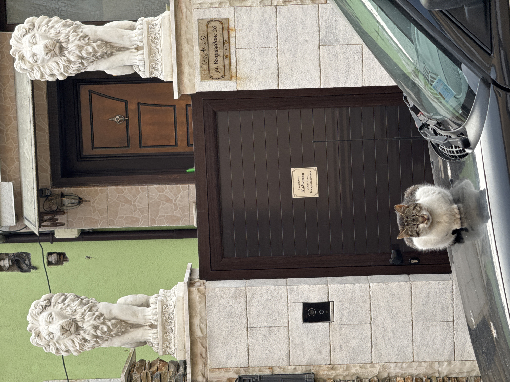 A residential scene features a two-story building facade in Burgas, Bulgaria. Two brown entry doors are visible, flanked by stone lion statues atop pillars. A street sign with the address "ул. Възраждане 26" (Vazrazhdane Street 26) is affixed near the upper door. A cat is resting on the hood of a gray car parked near the lower door. A sign on the lower door reads "Семейство Хаджиеви Иван, Анита, Тодор, Валентин" (Hadjievi Family: Ivan, Anita, Todor, Valentin). The building is painted with a lime green exterior.