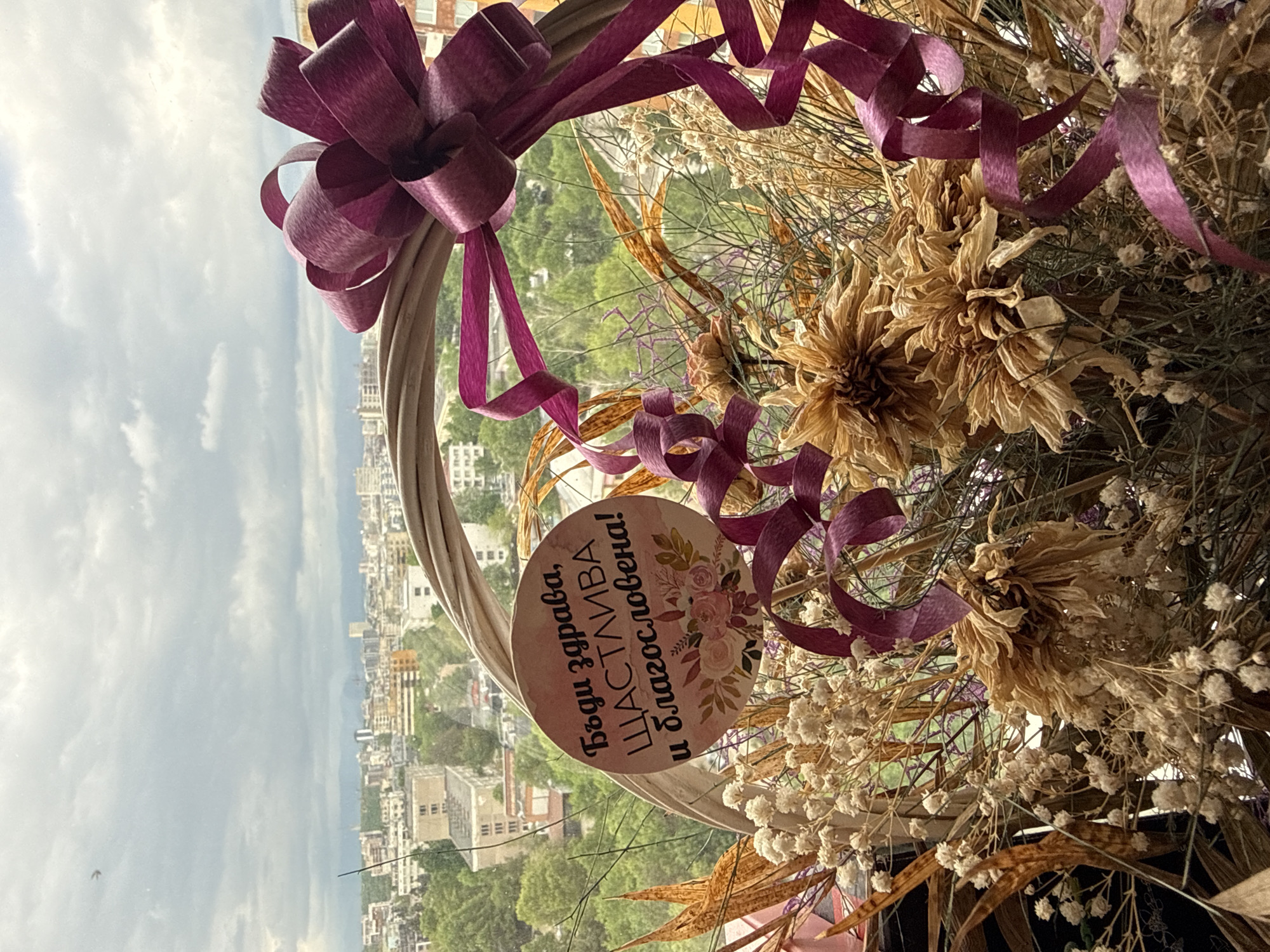 An eye-level, close-up shot displays a decorative wreath against an urban background, which is identifiable as the city of Sofia, Bulgaria.
The wreath, constructed from woven, natural materials, is adorned with dried flowers in shades of beige and white. A purple ribbon, tied in a bow, and additional spiraled strands of the same ribbon embellish the wreath. A circular card is attached to the wreath, bearing Bulgarian text and small floral illustrations.
The urban backdrop visible through the wreath includes buildings, trees, and the sky. The buildings vary in height and architectural style. The sky is overcast.