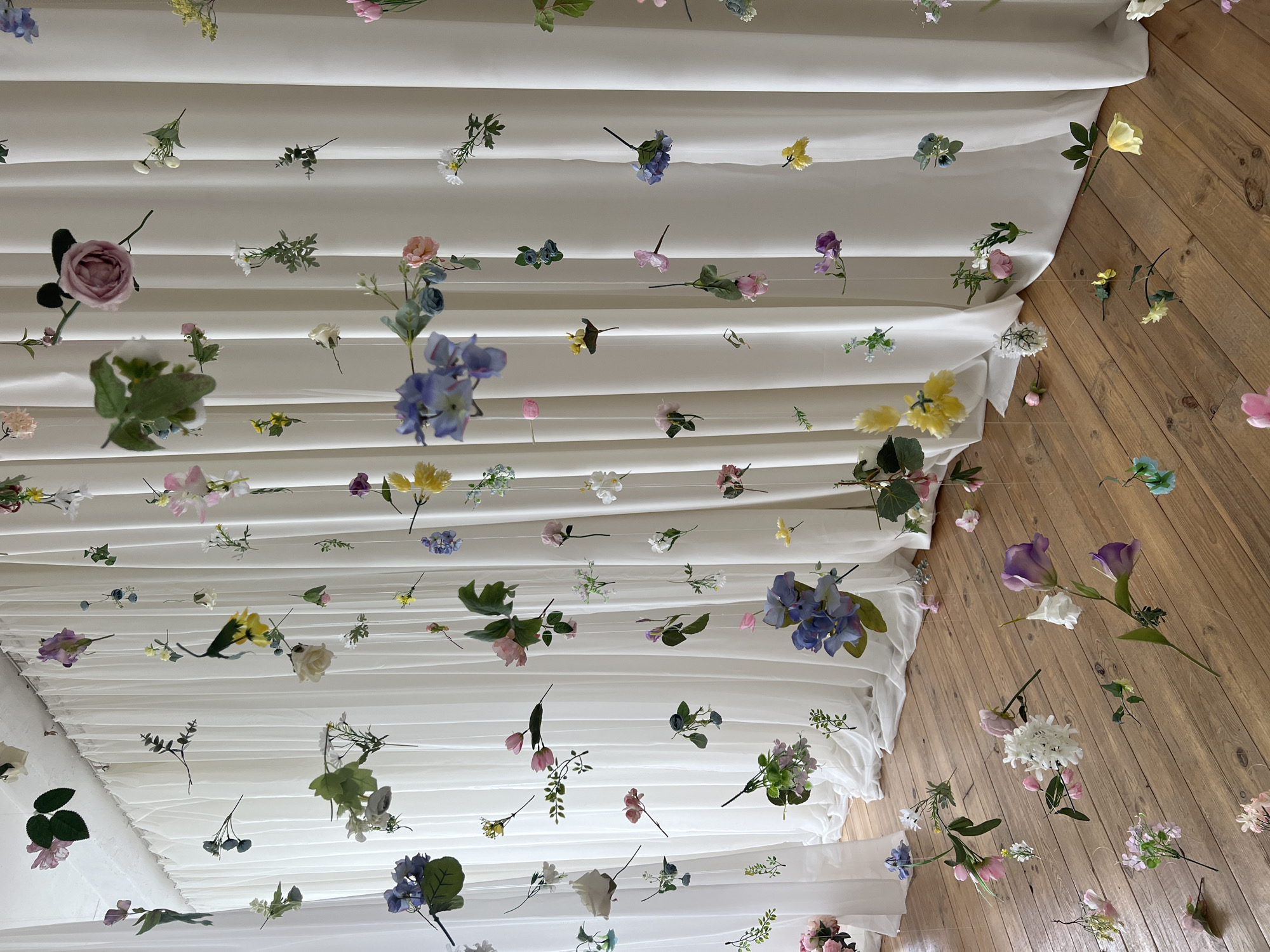 The image displays a whimsical arrangement of artificial flowers suspended from the ceiling by clear threads, creating a delicate, ethereal effect. The backdrop consists of floor-to-ceiling white curtains that drape gracefully, providing a soft, neutral canvas for the vibrant floral display. The floor is made of polished wooden planks, adding a warm, natural element to the scene.
The flowers themselves are diverse in color and form, with hues ranging from soft pinks and purples to bright yellows and whites. Some flowers are clustered together, while others hang individually, creating a dynamic and visually interesting composition. The lighting is bright and diffused, suggesting an indoor setting, possibly a studio or a well-lit room.
There are no people present in the image, and the focus is solely on the artistic installation of the flowers. The overall impression is one of lightness, beauty, and a touch of fantasy. The scene does not provide any specific visual cues that would indicate its location within the city of Kyiv, Ukraine.