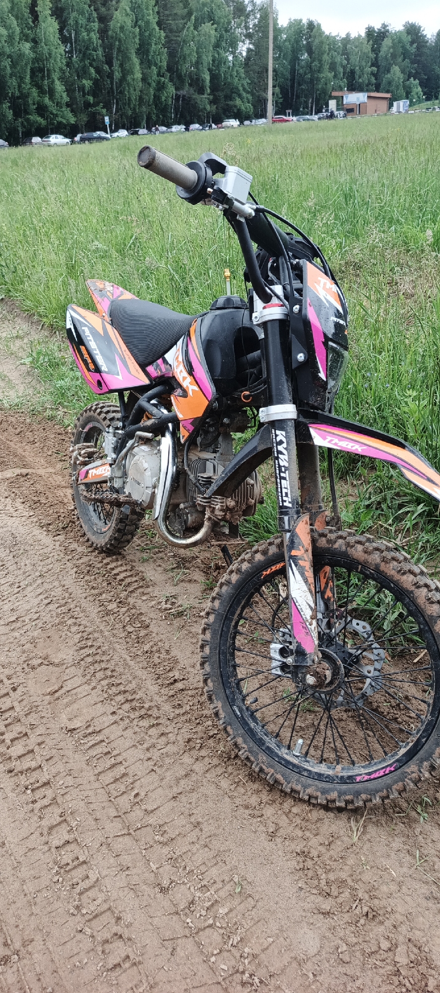 A heavily mud-splattered dirt bike, primarily black with orange and pink graphics, is parked on a dirt track in Прикамский, Russia. The bike features "PITSTER SPX," "MBK," "TMBK," and "KYB-TECH" branding on various components. Its knobby tires are covered in mud, as are parts of the engine, frame, and suspension. Distinct tire tracks are imprinted in the wet, reddish-brown soil beneath and around the bike. To the left and right of the track, tall green grass extends into the background. A dense line of trees forms a natural barrier in the mid-ground. Beyond the trees, a row of parked vehicles is visible, suggesting a nearby road or parking lot. A tall, slender pole stands amidst the trees, and a small brown building is partially visible in the distant background to the right. The sky appears overcast. No individuals are present in the scene.
