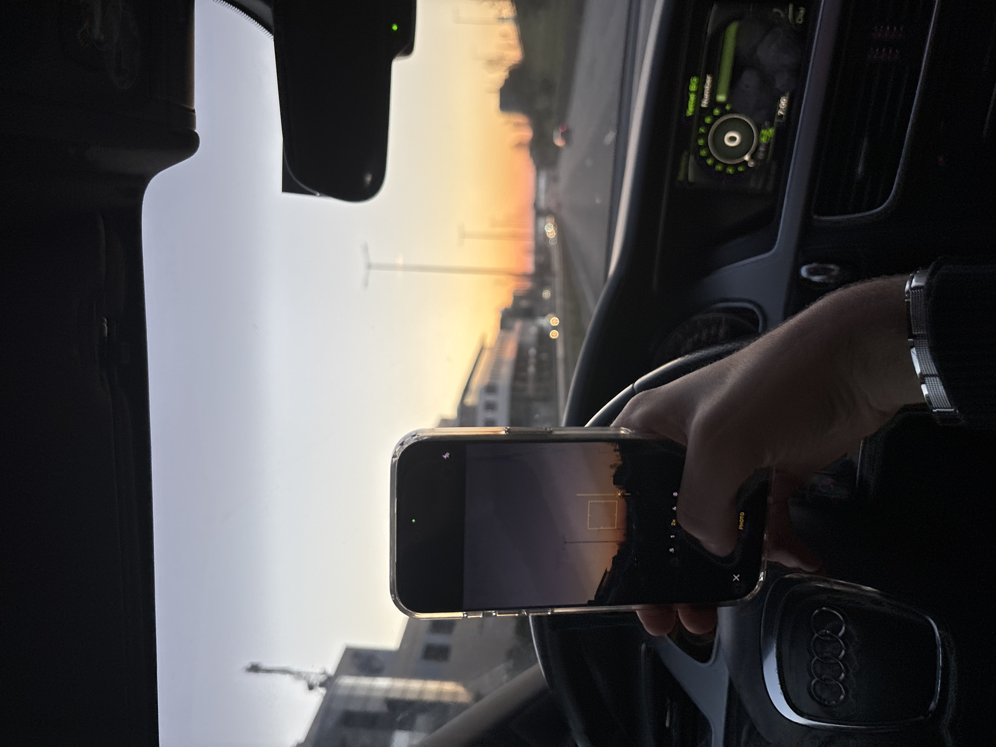 The scene is a person taking a photo with their smartphone, inside a vehicle, possibly an Audi, while driving on a road during sunrise or sunset. The car's dashboard and steering wheel are visible. The phone's screen shows the image being captured of the outdoor scenery which includes street lights and buildings. The time on the car's dashboard shows 7:00. Location: Likely on a highway or major road in Sofia, Bulgaria.