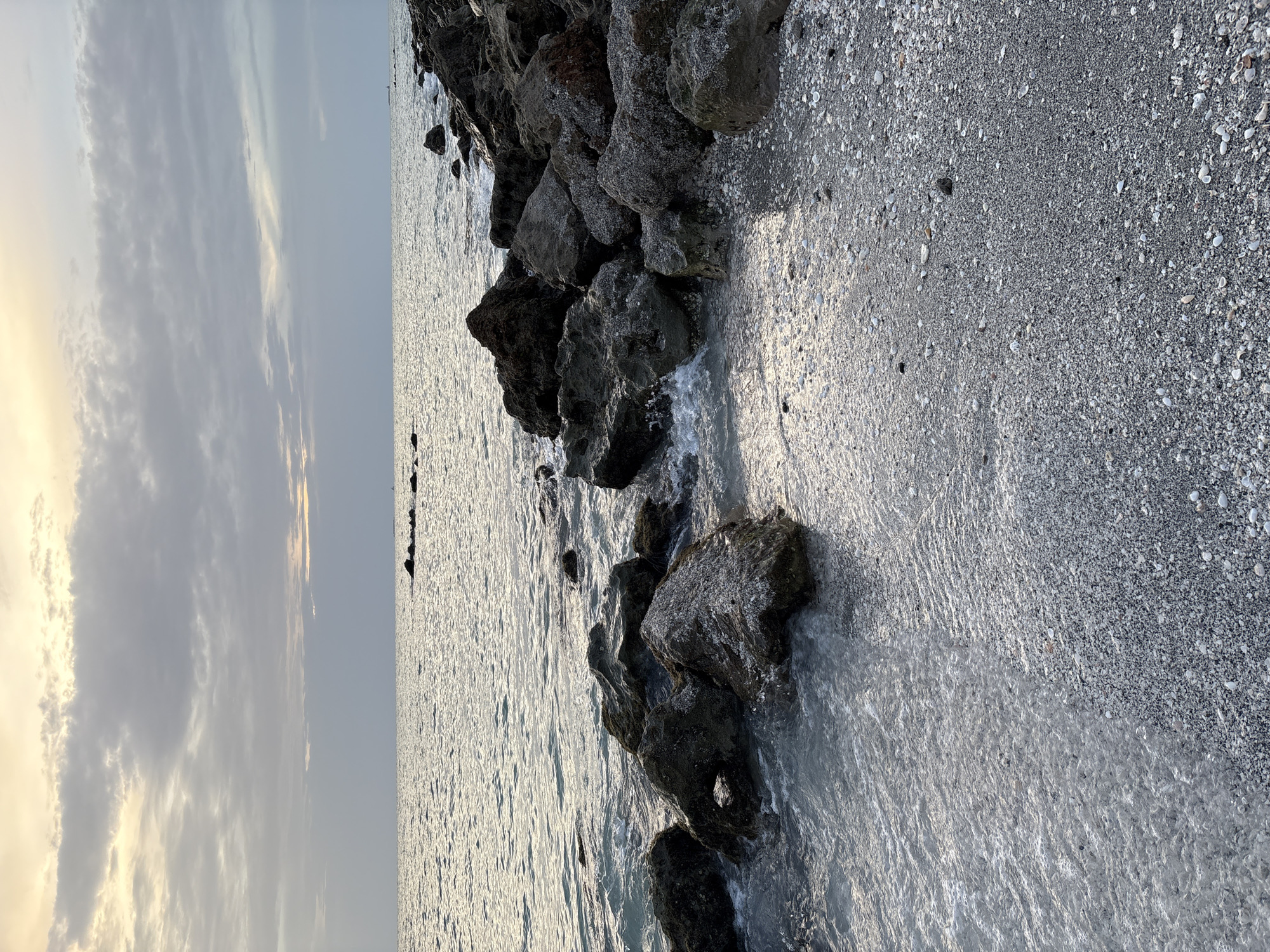 The image shows a rocky beach at sunset. The sky is cloudy, with the sun setting behind the clouds, casting a warm glow. The ocean is calm, with gentle waves lapping at the shore. The beach is covered in small stones and shells, and there are several large rocks scattered along the waterline. In the distance, a small lighthouse is visible. The location is Venice, Florida, USA.
