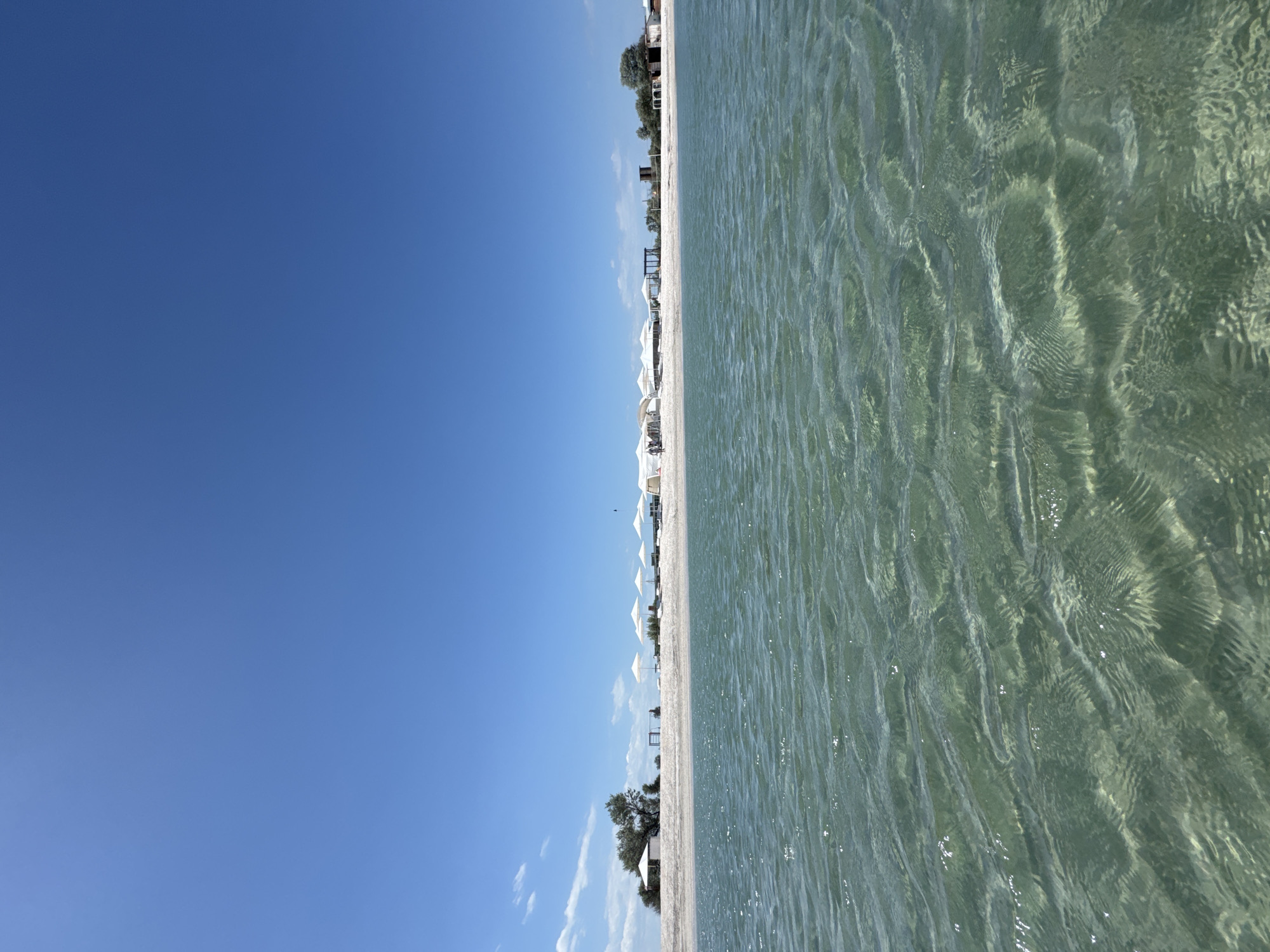 The image depicts a beach scene in an unknown location. The sky is a clear, bright blue. The water is clear and shallow, with visible ripples and patterns on the surface. A sandy beach separates the sky and water, with several white beach umbrellas and occasional trees along the coastline. The horizon is straight and clearly defined. There are no people visible in the scene.