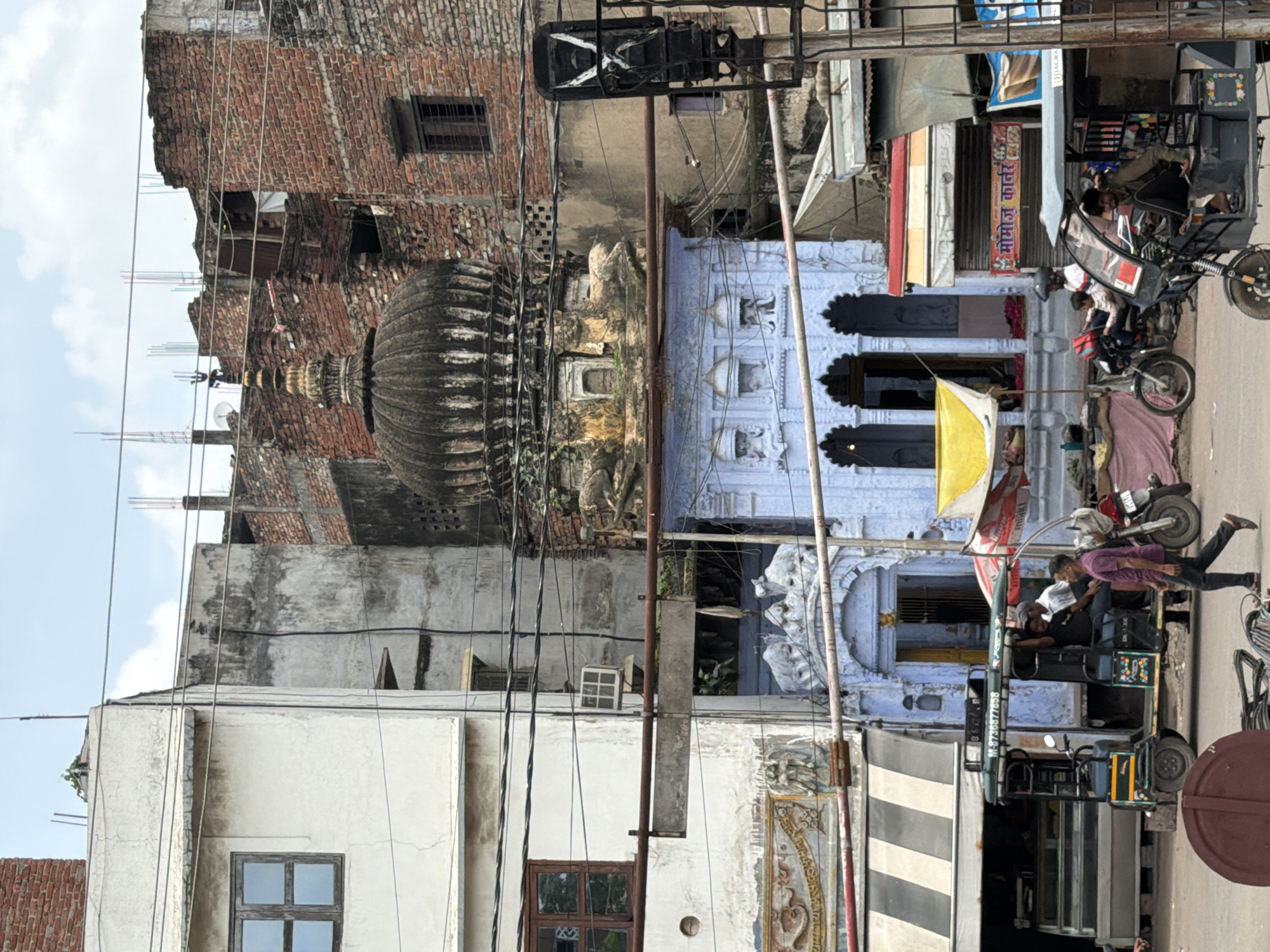This is a street-level, eye-level, medium shot captured in Kanpur, India, likely in a bustling market area. The scene is dominated by a variety of buildings and vehicles.
In the foreground, a maroon-colored circular object rests on the ground, possibly a large lid or a decorative element. To its right, a pedestrian is walking away from the camera, wearing a maroon t-shirt and dark pants. Behind them, an auto-rickshaw is parked, with several people inside and one standing near the entrance. To the left of the auto-rickshaw is a motorcycle with a rider and a passenger. Further left, a food stall with a striped awning covers a display case.
The middle ground features a light blue building with an ornate facade, featuring arched doorways and decorative sculptures. Above this building, a dark, domed structure, likely part of a temple or mosque, rises prominently. This domed structure is flanked by older, brick buildings with exposed construction elements and satellite dishes.
The background consists of more buildings constructed from brick and concrete, some appearing weathered and incomplete. Electrical wires are strung across the frame, suggesting an urban environment. The sky above is overcast with white clouds. The overall impression is of a dense urban environment with a mix of old and new architecture, and active street life. The presence of the auto-rickshaw, the clothing of the people, and the signage with Devanagari script strongly indicate a location in India, specifically Kanpur given the prompt.
