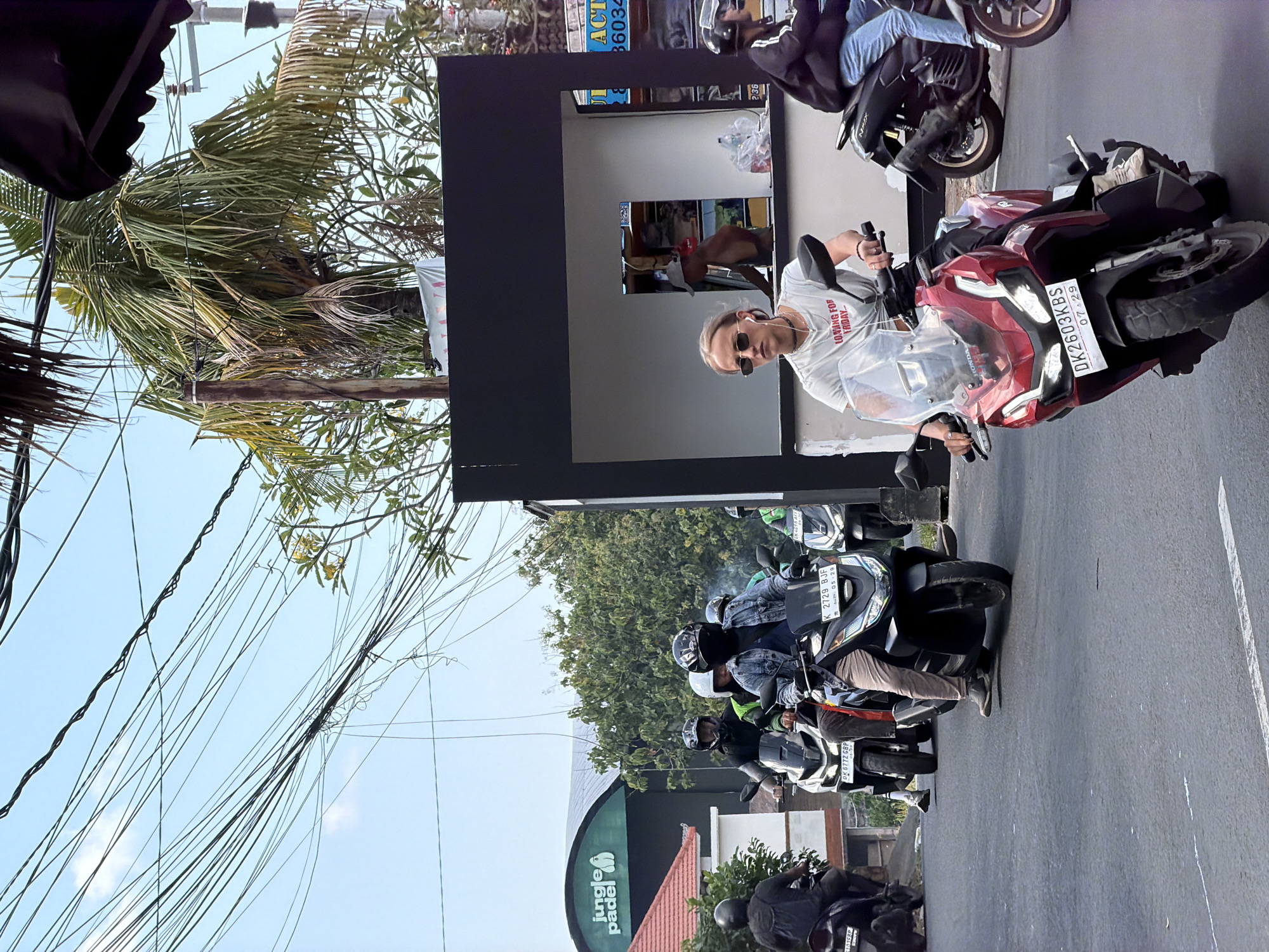 This image, rotated 90 degrees counter-clockwise, depicts a busy street scene in what appears to be North Kuta, Indonesia. The setting is bright daylight under a clear sky.
In the prominent foreground, a person wearing sunglasses and a white t-shirt with the text "LOMING FOR FRIDAY" (likely "LOOKING FOR FRIDAY") is seated on a red Honda motorcycle. The motorcycle bears the license plate DK 2603 KBS. This individual is looking towards the camera, gripping the handlebars. Behind them, another rider on a black scooter is partially visible.
Further into the background, a line of several more people on motorcycles, many wearing helmets, are visible on the road, suggesting a traffic queue or a momentary stop. Other visible license plates include K 2729 BJF and DK 6772 GBP.
The surroundings feature lush tropical vegetation, including prominent palm trees and other dense green foliage. Overhanging the street is a dense tangle of electrical wires against the blue sky. On the side of the road, various buildings and storefronts are present. A prominent sign for "Jungle Padel" is visible on a building, and another partially obscured sign shows "ACT 36034." The overall scene captures a typical vibrant street environment characterized by abundant motorcycles and tropical flora.