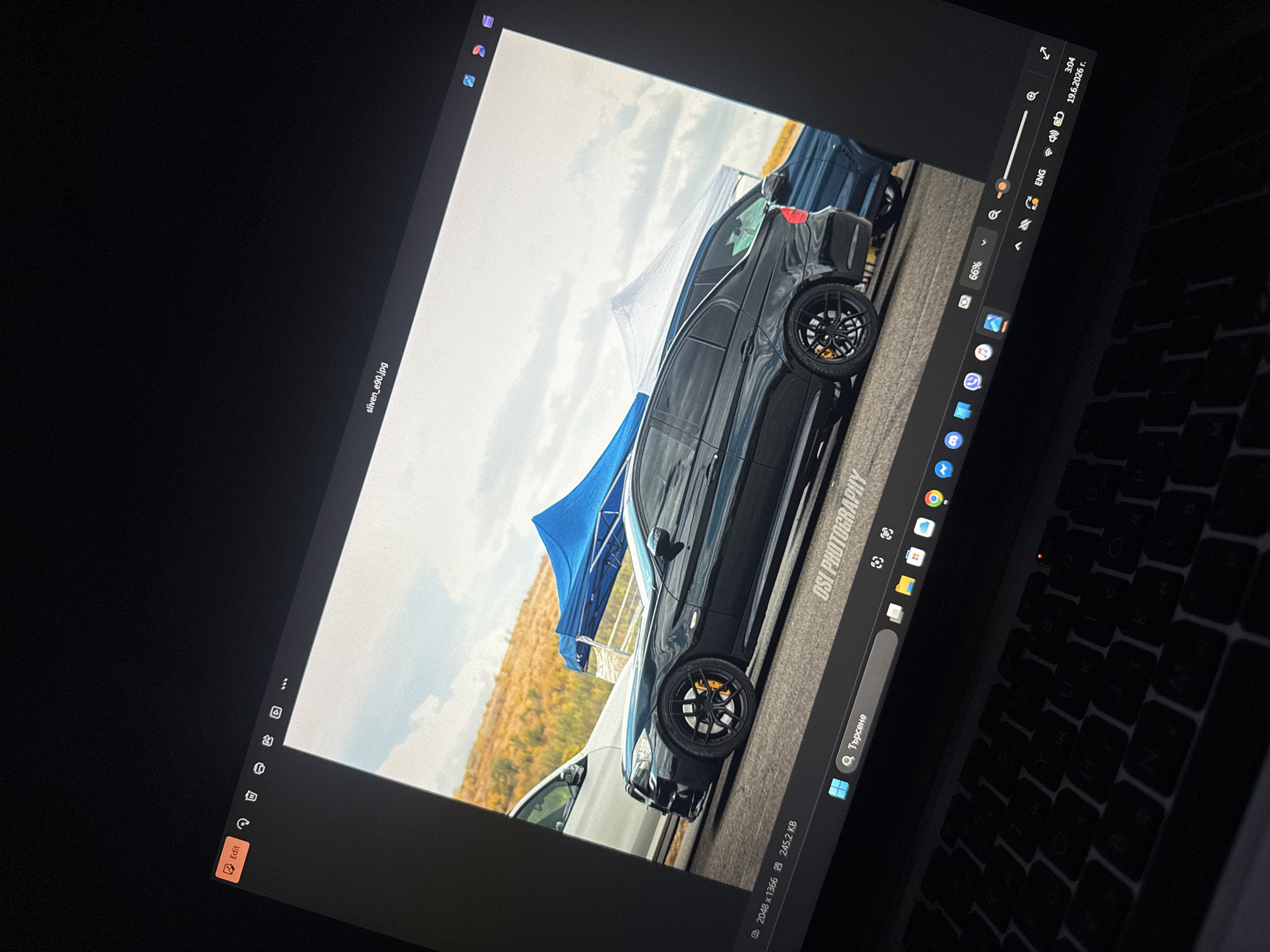 The image shows a laptop screen displaying a photograph of a black BMW wagon. The car is parked on a road with a dirt embankment covered in dry grass and trees in the background. Two canopies, one blue and one white, are set up behind the car. The laptop is open, and the keyboard is visible in the foreground. The operating system appears to be Windows, with a taskbar displaying icons for various applications and system functions. A file name "sliven_e90.jpg" is visible in the top right corner of the image displayed on the screen. The timestamp "3:04" and date "19.6.2026 r." are also visible in the lower right corner of the taskbar. The term "Dimitrovgrad, Bulgaria" is not present in the image, nor is there any visual information to suggest the location.