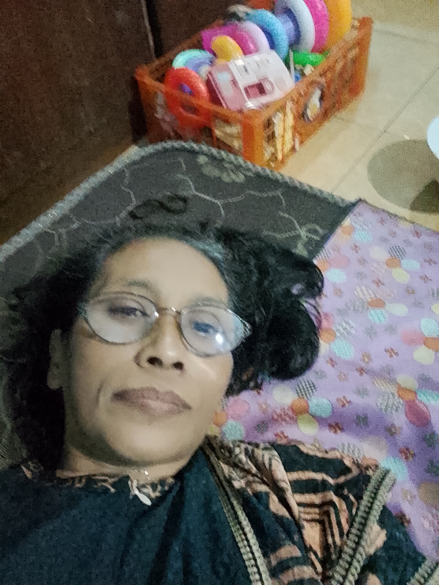 In the city of Kecamatan Pati, Indonesia, an overhead, selfie-style shot captures a woman lying down on a purple, floral-patterned blanket. She is wearing glasses and a dark shirt with a patterned shawl draped over it. To her right, on a dark patterned mat, is an orange plastic basket filled with colorful children's toys, including rings of various colors and a pink toy house. The background is dimly lit, with a dark brown textured surface to the left and a lighter, textured floor to the right.