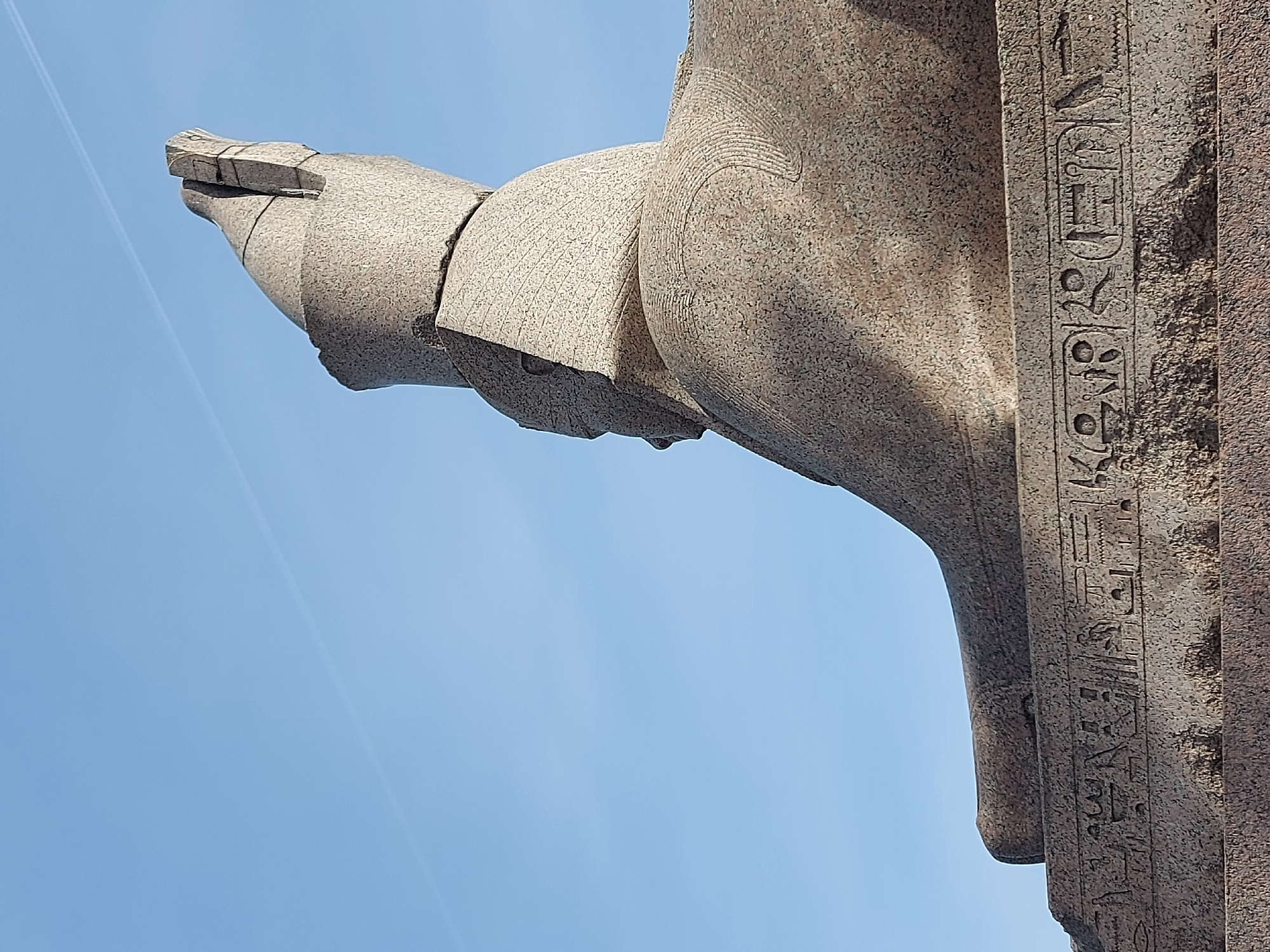 The image shows a close-up of a granite Egyptian sphinx statue against a clear blue sky. The statue appears to be weathered and slightly damaged, with visible cracks and chips. Hieroglyphic inscriptions are present on the side of the statue. The sphinx is located on the Universitetskaya Embankment of Saint Petersburg, Russia. The angle suggests the photo was taken from a low perspective, looking upwards at the statue.