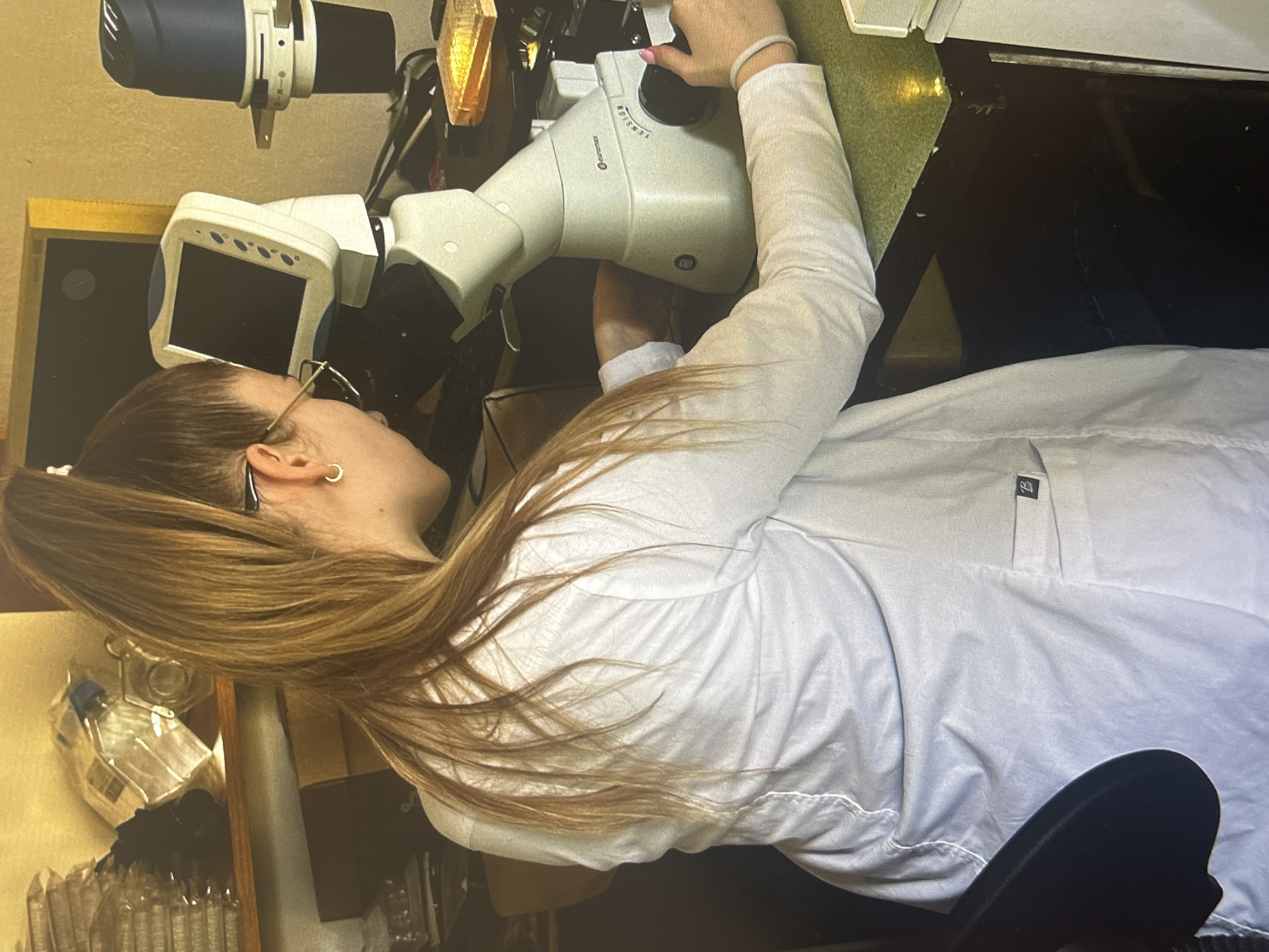 A woman with long blonde hair pulled into a ponytail is looking through a microscope. She wears glasses and a white lab coat. The microscope is a complex piece of equipment with a screen and various components. The scene appears to be a laboratory setting. The city is unknown.