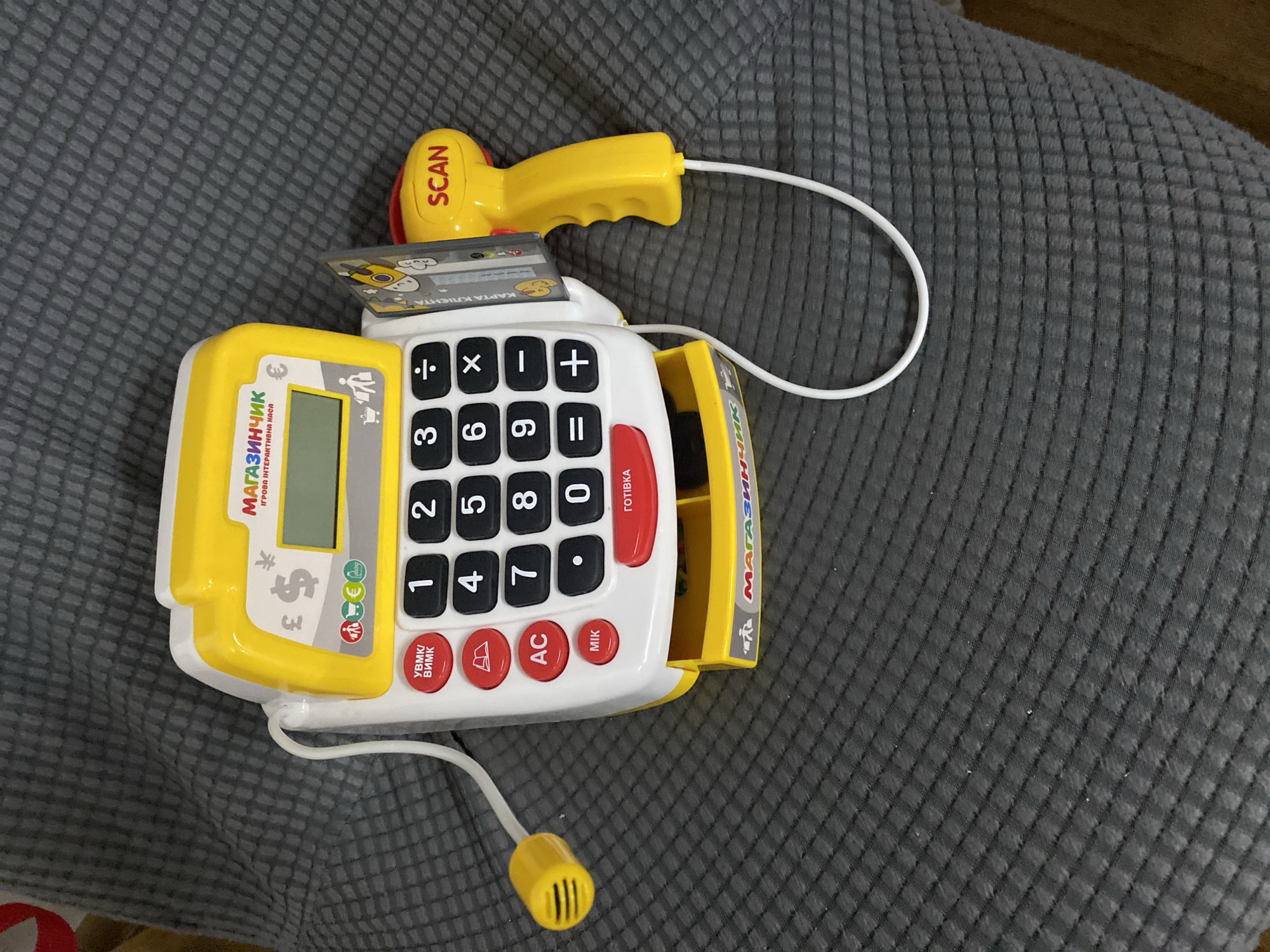 This image shows a toy cash register and scanner placed on a dark, textured surface, likely a piece of furniture or fabric. The cash register is predominantly yellow and white, with a keypad displaying numbers 0-9 and arithmetic symbols. It also features red buttons labeled "УВМК/ВИМК", "AC", "MIK", and a larger red button labeled "ГОТІВКА". A small green display screen is visible at the top of the register, showing a coin and a shopping cart icon. Above the screen, the text "МАГАЗИНЧИК" is printed in red, followed by "ІГРОВА ІНТЕРАКТИВНА КАСА" in smaller black text. There are also currency symbols like £, ¥, and € displayed on the front of the register.
A yellow scanner gun with the word "SCAN" in red is attached to the side of the cash register by a white cord. A toy credit card is inserted into a slot on the cash register. The draw for the cash register is partially pulled out, revealing a green object inside. The background is a dark gray, quilted texture.
There is no indication or information in the image that places this scene in Kryvyi Rih, Ukraine.