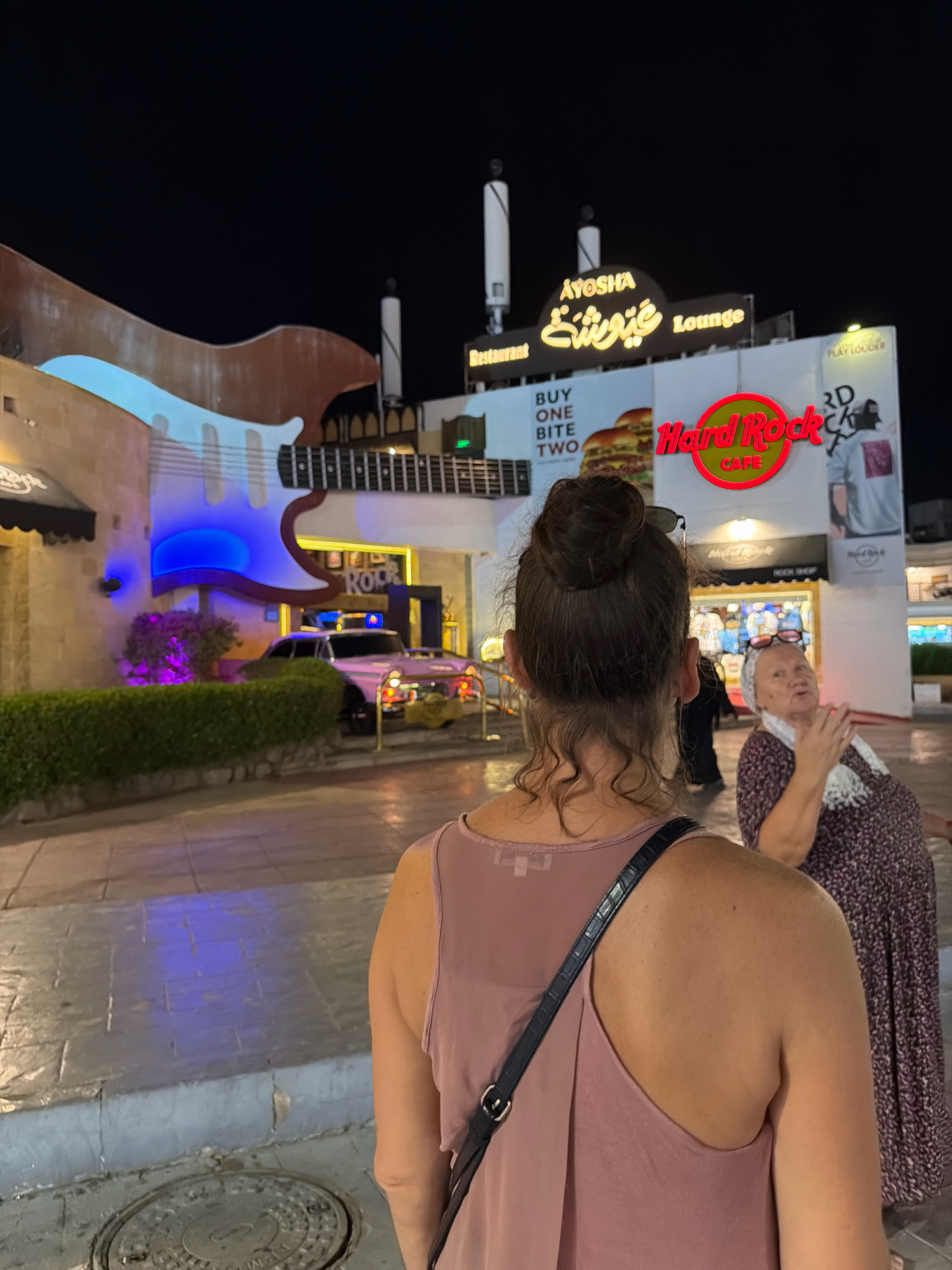 A medium shot captures two women in the foreground, facing away from the camera, in front of a Hard Rock Cafe in Sharm El Sheikh, Egypt, at night. The woman on the left has her brown hair pulled up in a bun. She is wearing a pink sleeveless top. The woman on the right is wearing a patterned dress and a white scarf around her neck. She is looking towards the left with her mouth open, as if speaking. In the background, a large replica of a guitar is mounted on the building, illuminated with blue lights. A sign above the entrance reads "AYOSHA Restaurant Lounge" in English and Arabic. The Hard Rock Cafe logo is prominent on the building. A pink vintage car is parked in front of the establishment, and a hedge is visible. The ground is paved with stone tiles, and a manhole cover is in the lower left corner. The overall scene suggests a lively tourist area at night.