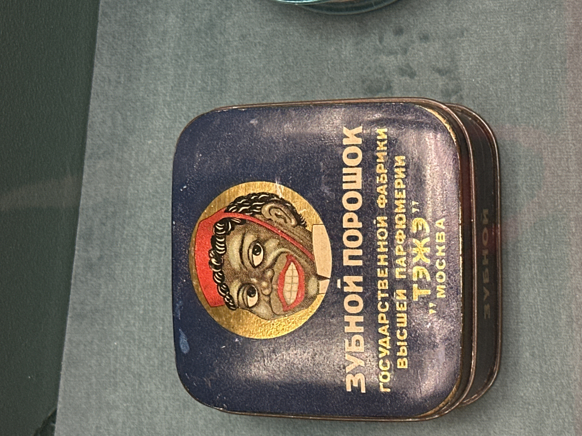 The image shows a close-up, eye-level shot of a vintage tin of "ZUBNOY POROSHOK" (tooth powder) produced by the "GOSUDARSTVENNAYA FABRIKA VYSSEI PARFYUMERII" (State Factory of High Perfumery) in Moscow. The tin is dark blue with gold lettering. A circular emblem on the lid features a cartoonish illustration of a Black man wearing a red fez and a white shirt, with a wide smile and prominent teeth. The text below the emblem reads "GOSUDARSTVENNOI VYSSHEI PARFYUMERII TЭЖЭ" and "MOSKVA." The tin is resting on a textured, teal-colored surface, possibly a display cloth or a shelf. The lighting is neutral, and the focus is sharp on the tin.