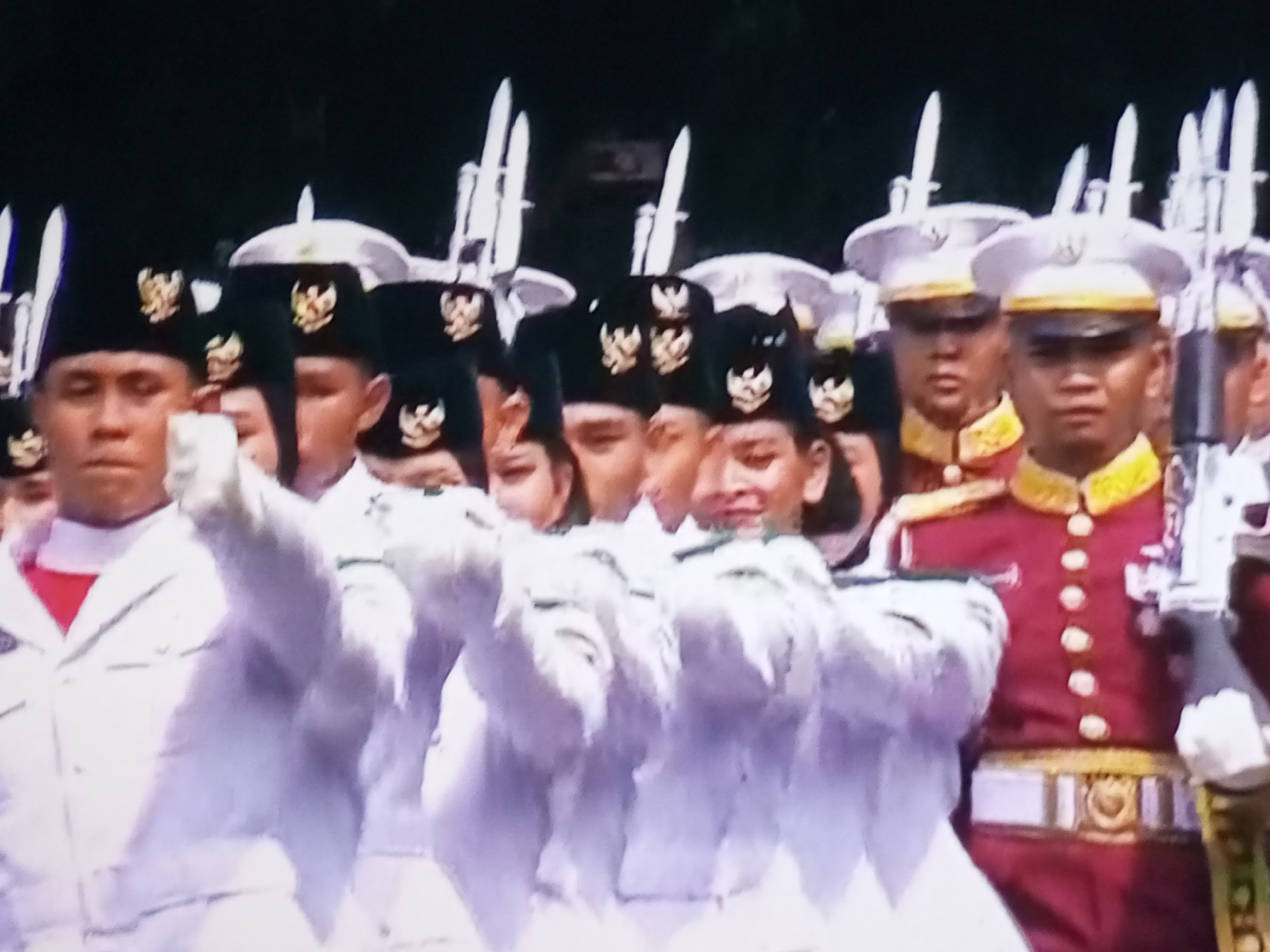 The image shows a contingent of soldiers marching in formation. They are wearing dark caps with a crest, white shirts, and dark jackets with gold trim. Some soldiers are holding rifles with bayonets. The soldiers appear to be marching in a parade or ceremony. The background is dark, suggesting it might be an outdoor event. There is no clear indication of the location being Banyudono, Indonesia, as there are no identifiable landmarks or text in the image to support this claim.