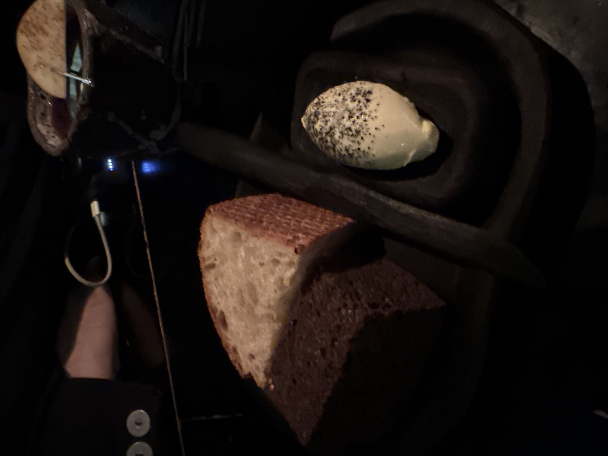 The image captures a dimly lit, culinary still life, likely in a restaurant. Visible are pieces of food presented in a dark and somewhat abstract arrangement. A slice of bread, partially white and partially dark, sits prominently in the lower half of the frame. Above and to the right is a small bowl, possibly made of dark clay or ceramic, containing a dollop of white cream topped with black specks. To the upper left are other pieces of food, possibly skewered on a thin metal rod. The overall darkness and composition suggest a sophisticated, modern culinary style. Given the nature of the food and presentation, it’s difficult to definitively determine that this scene is specifically located in Moscow, Russia without additional context.