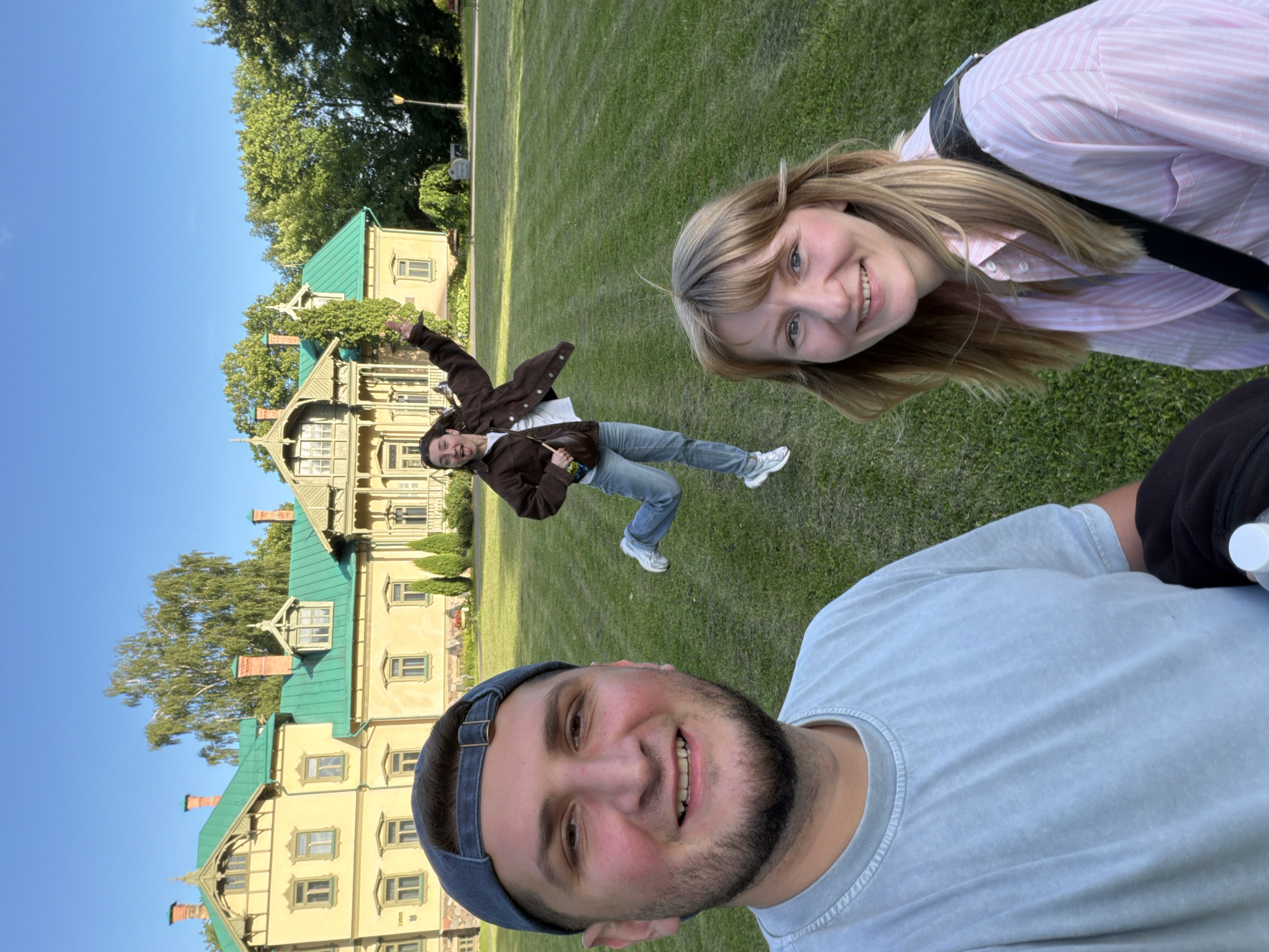 Three people are posing for a selfie in front of a light yellow building with green trim and a green roof. The building appears to be a historic estate or manor house. A man in a light blue t-shirt and a blue baseball cap is in the foreground, smiling at the camera. To his right and slightly behind him is a woman in a pink and white striped shirt, also smiling. In the background, a third person, a woman wearing a brown jacket over a white shirt and blue jeans, is captured mid-jump with one arm raised and her tongue sticking out. The ground is covered in green grass, and trees are visible in the background. The sky is clear and blue. The location appears to be the manor complex of the Belarusian State Agricultural Academy in Gorki, Mogilev Region, Belarus, not Minsk.