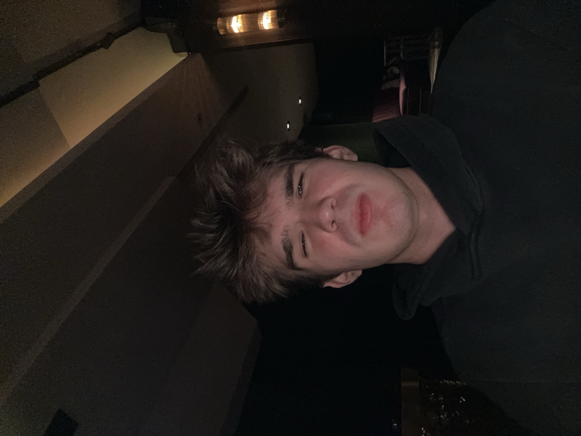 The image captures a young adult male from the chest up, centered in a dimly lit indoor environment. He has light brown, tousled hair and fair skin. His eyes are tightly squinted, and his mouth is slightly open with lips appearing somewhat pursed or swollen, giving his face an expression of discomfort or grimace. He is wearing a dark, possibly black, hooded sweatshirt, with the hood resting as a thick collar around his neck.
The background is dark and suggestive of a bar, lounge, or upscale restaurant in Warsaw, Poland. To the right of the subject, a wall features a tall, cylindrical wall sconce emitting a warm, amber light, possibly with multiple tiers of illuminated glass or translucent material. Further behind, a dark red or maroon upholstered chair with a high back is partially visible, along with what appears to be a dark table or stand. The ceiling shows architectural elements like beams or panels, with some areas lit by warm ambient light. Two small, circular, recessed lights are discernible in the upper right portion of the ceiling or a darker panel. The left side of the background is largely obscured by shadow but shows a dark, textured surface, possibly a wall or paneling. The overall lighting scheme creates a cozy yet subdued atmosphere.