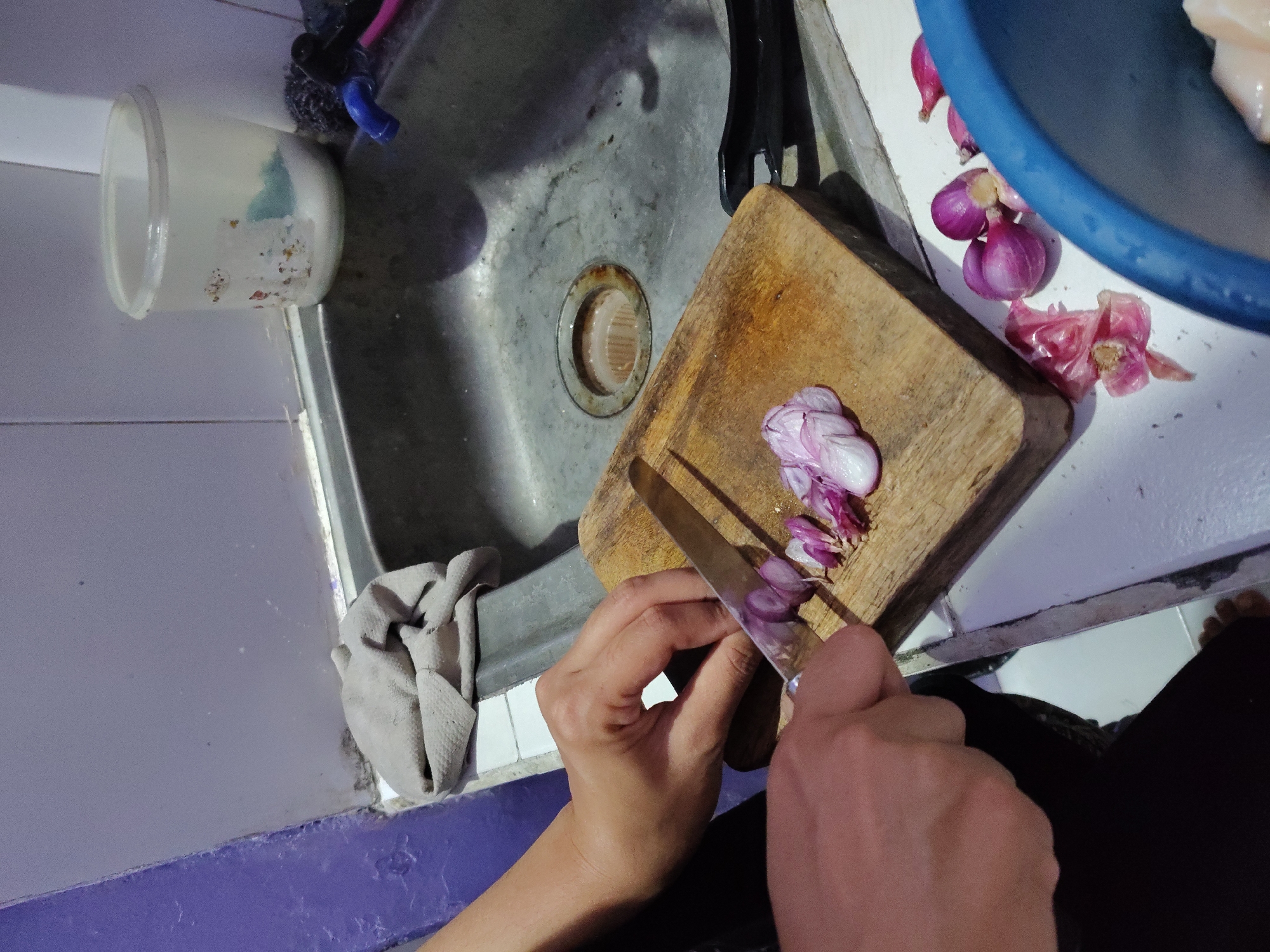 A medium shot captures a person's hands in the process of chopping red onions on a wooden cutting board. The cutting board is positioned on a white countertop next to a stainless steel sink. Several red onions, some whole and some peeled and chopped, are scattered on the countertop and the cutting board. The sink contains a drain with a metal grate and some debris. To the right of the sink, a blue plastic basin is partially visible, containing what appears to be raw chicken. In the background, a white plastic container with a lid, possibly for storage or hygiene products, sits on the edge of the sink. A gray cloth is draped over the edge of the countertop near the sink. The walls surrounding the sink are tiled in a light purple color. The lighting suggests an indoor environment, likely a kitchen. This scene is consistent with food preparation activities observed in residential kitchens across Surabaya, Indonesia, where such common household items and cooking practices are prevalent.