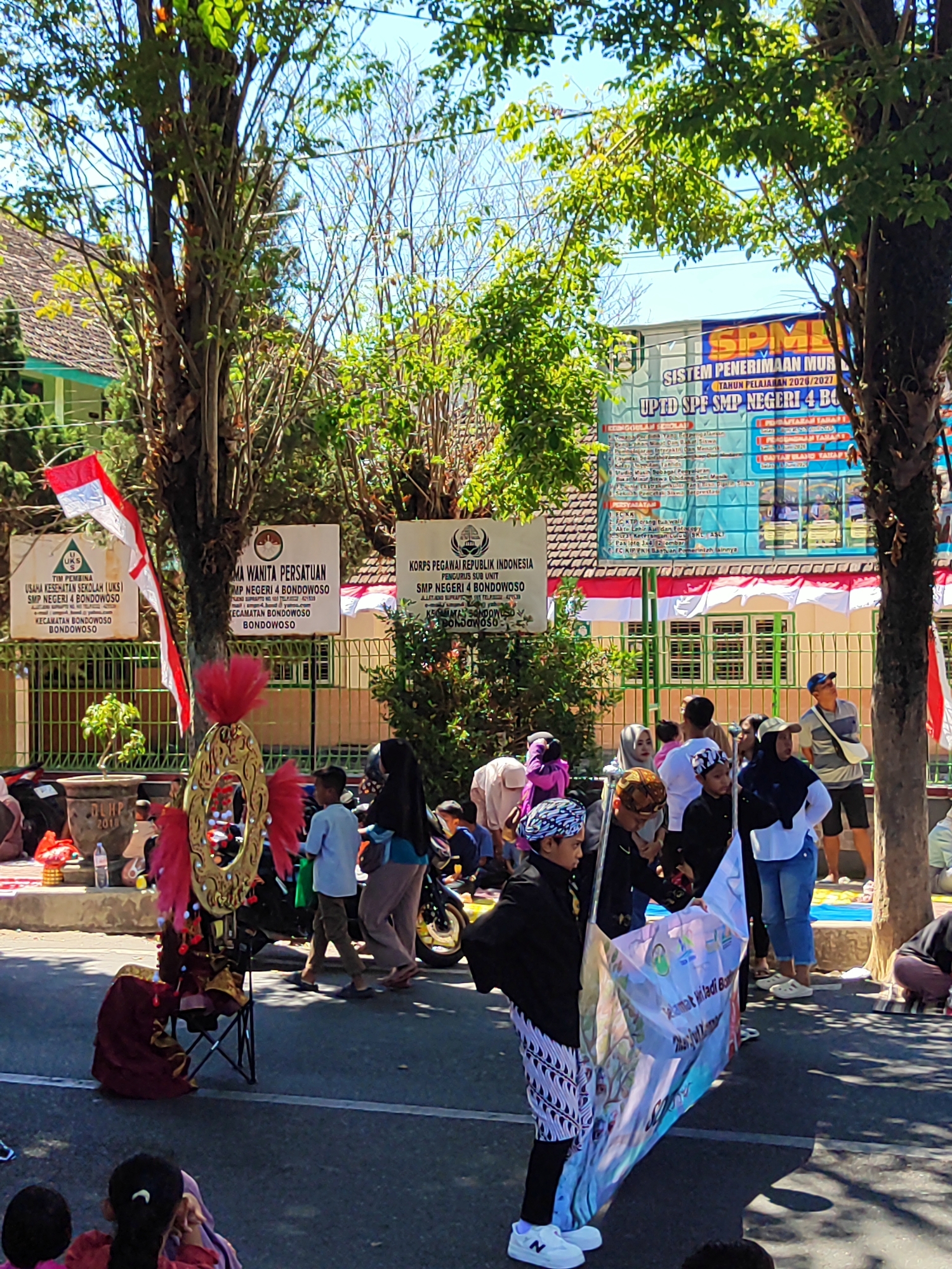 The image depicts an outdoor event in Dabasah, Indonesia, possibly a school or community gathering. In the foreground, a person in traditional Javanese attire, including a batik sarong and headcloth, holds a large banner. Another person, dressed in a maroon and gold costume with feathered headwear, sits on a foldable chair next to a decorative stand. Several other individuals, including children and adults, are visible in the midground and background, some observing the event and others participating. Behind them, a building with a green fence and white banners hangs in front of it, suggesting an institutional setting. Indonesian flags are prominently displayed, indicating a national celebration or patriotic event. The scene is illuminated by bright sunlight, casting shadows on the pavement, and lush green trees provide shade. The overall atmosphere appears festive and communal.