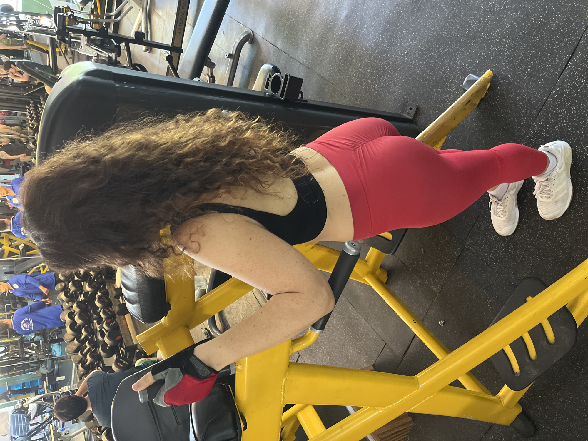 A medium shot captures a woman with long, curly brown hair wearing a black sports bra and red leggings. She is positioned on a yellow weight machine in a gym. Her right hand, adorned with a red and black weightlifting glove, is on the machine's handle. Her left arm is bent, with her elbow resting on the padded part of the machine. Her feet are clad in white athletic shoes with white socks. The gym environment features weight racks filled with dumbbells in the background, other gym equipment, and a black rubberized floor. The overall scene is within a fitness facility. The image does not contain any specific indicators of location within Ham Thang Ward, Vietnam.