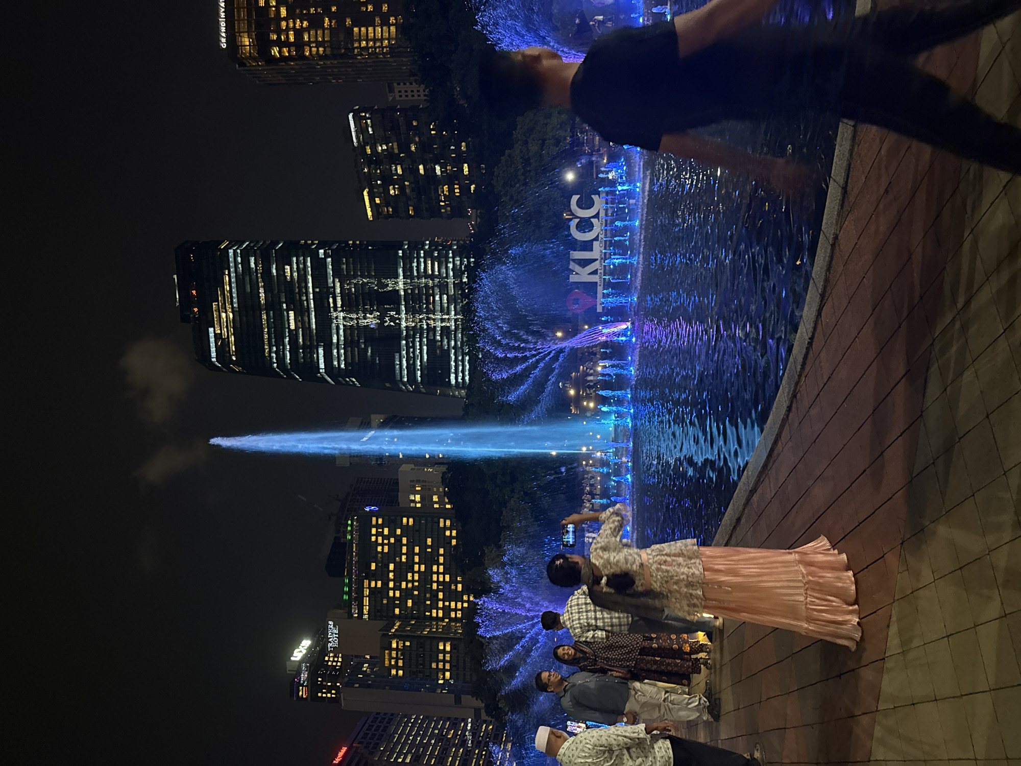 The image depicts a nighttime scene in Kuala Lumpur, Malaysia, likely near the Petronas Twin Towers. A large, illuminated fountain with the letters "KLCC" is prominently displayed. A group of people are standing near the fountain, some of whom are taking pictures with their phones. In the background, there are illuminated skyscrapers and other buildings. A person in dark clothing is partially visible in the upper right corner of the image.