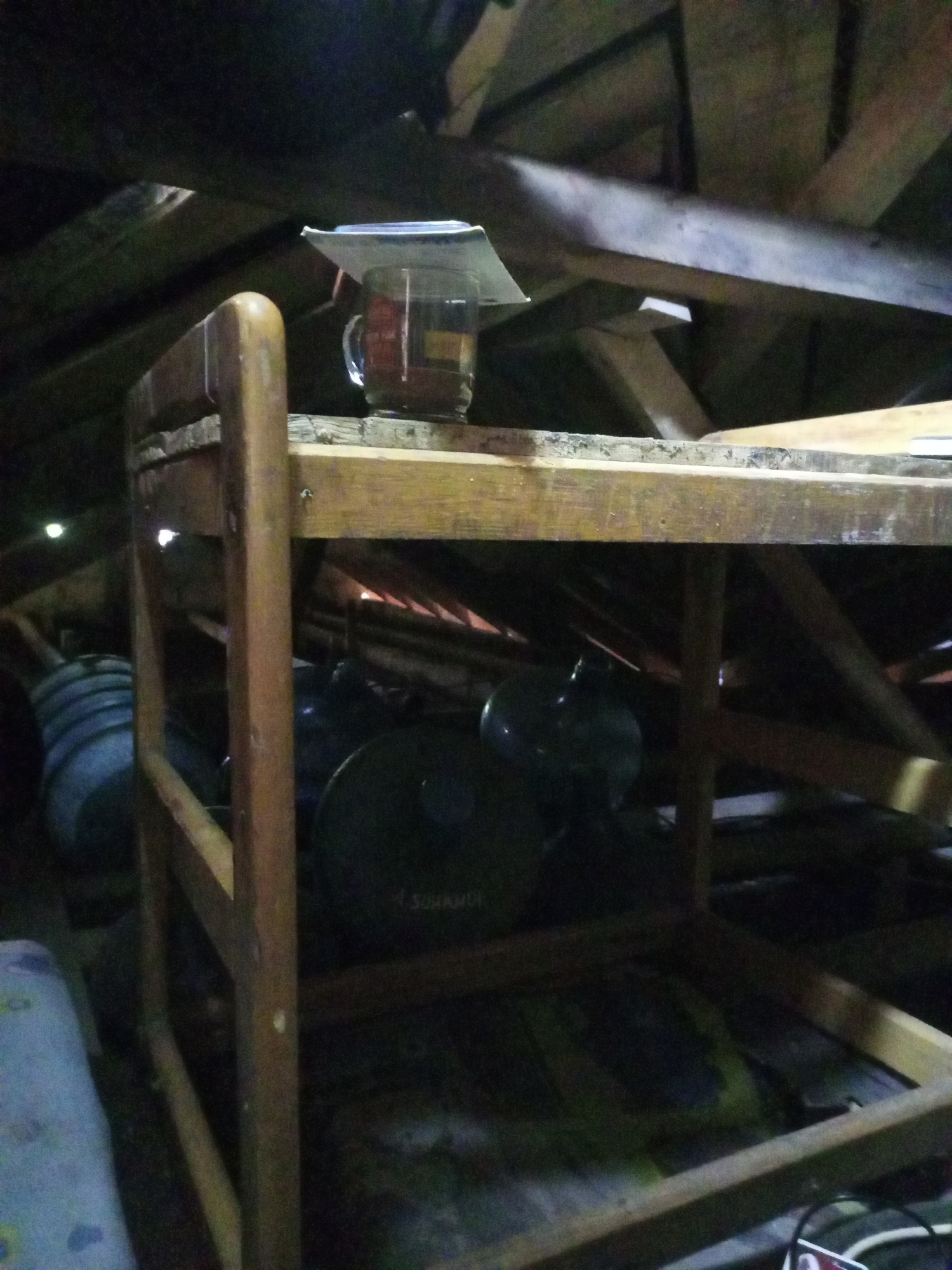 The image depicts the interior of an attic space, likely in a residential structure located in Cidahu, Indonesia. The attic features exposed wooden beams and rafters, typical of traditional Indonesian construction. A rudimentary wooden bed frame, seemingly constructed from unfinished lumber, is centrally positioned. On the upper level of this bed frame, a glass mug containing a dark liquid, possibly tea or coffee, is placed. The mug is partially covered by a piece of white paper or thin cardboard. Below the bed frame, several large, dark-colored cylindrical objects are visible, one of which appears to be a water dispenser. The overall impression is one of a storage or utility area, dimly lit and somewhat cluttered.