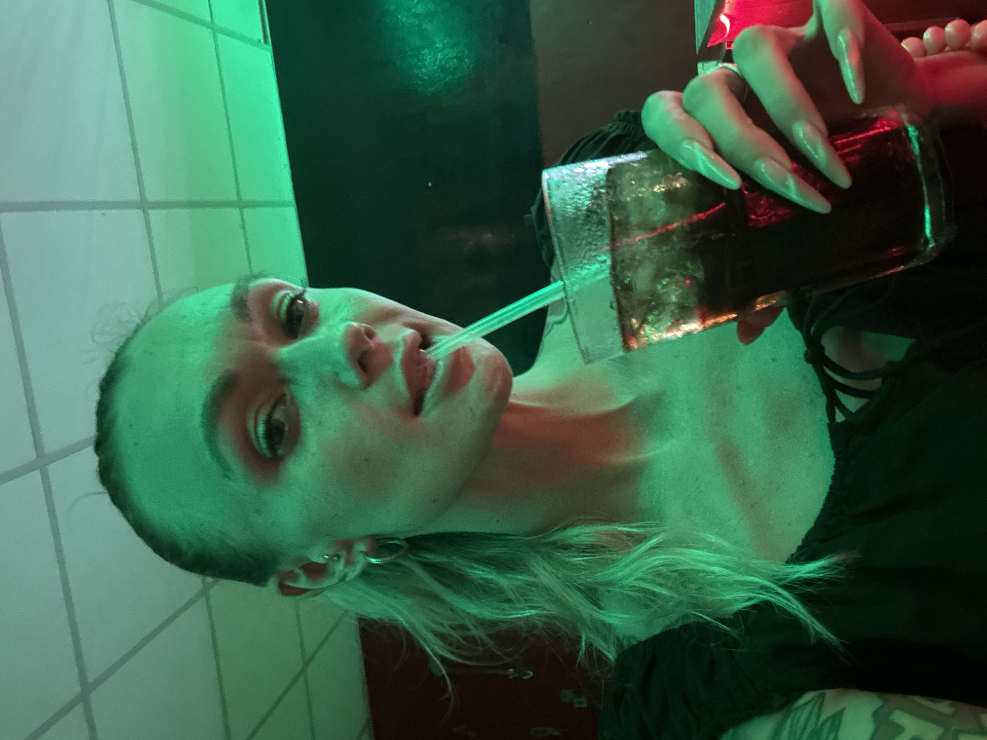 A woman with platinum blonde hair is holding a glass of dark liquid, possibly cola, with ice and a green straw to her lips. She is wearing a black top. Her fingernails are long and pointed. The background is illuminated by green and red neon lighting, with a tiled wall visible on the left. The location is possibly a bar or nightclub. The city is Mũi Né, Vietnam.