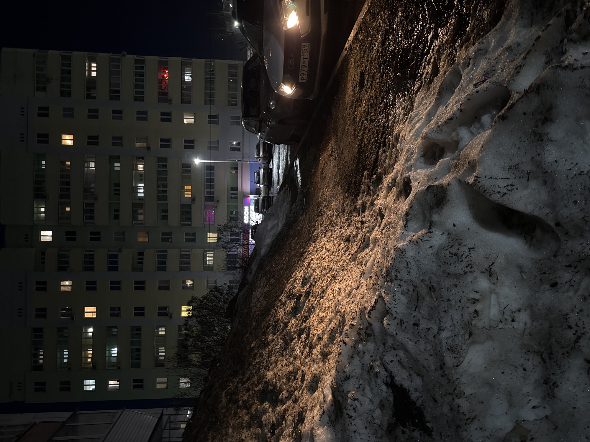 The image captures a nighttime scene in the city of Chaikovsky, Russia, during what appears to be late winter or early spring, characterized by melting snow. The foreground is dominated by a large, irregular bank of dirty, partially melted snow and ice, exhibiting footprints and dark patches of debris.
To the right of the snowbank, a wet, dark asphalt surface, indicative of a street or parking area, extends into the midground. Several vehicles are parked along this surface. The closest vehicle, positioned on the right, is a light-colored sedan (likely white or silver) with its headlights illuminated, casting light onto the ground. Its front license plate, visible and legible, reads "P979PT 159," which is a Russian Federation plate. Behind this car, a darker sedan is parked, and additional vehicles are visible further down the street.
A tall streetlamp with a bright white light illuminates parts of the street and the side of a building in the mid-background. Bare bushes or trees are situated between the snowbank and the buildings.
The background is dominated by a multi-story residential building, likely an apartment block, with a light-colored facade (possibly pale green or beige). Numerous windows are visible across its many floors; a significant number are illuminated with various hues, including white, yellow, and orange, indicating interior lighting. Notably, one window on an upper-mid floor exhibits a distinct red light, and another on a lower floor shows a purple light. Faintly visible signs, including one that reads "OZON" and another "KOTAK," are present on the ground-level facades of structures in the mid-background. No people are visible, and no direct actions or interactions are depicted. The scene is illuminated by artificial light sources from the streetlamp and building interiors, set against a dark night sky.