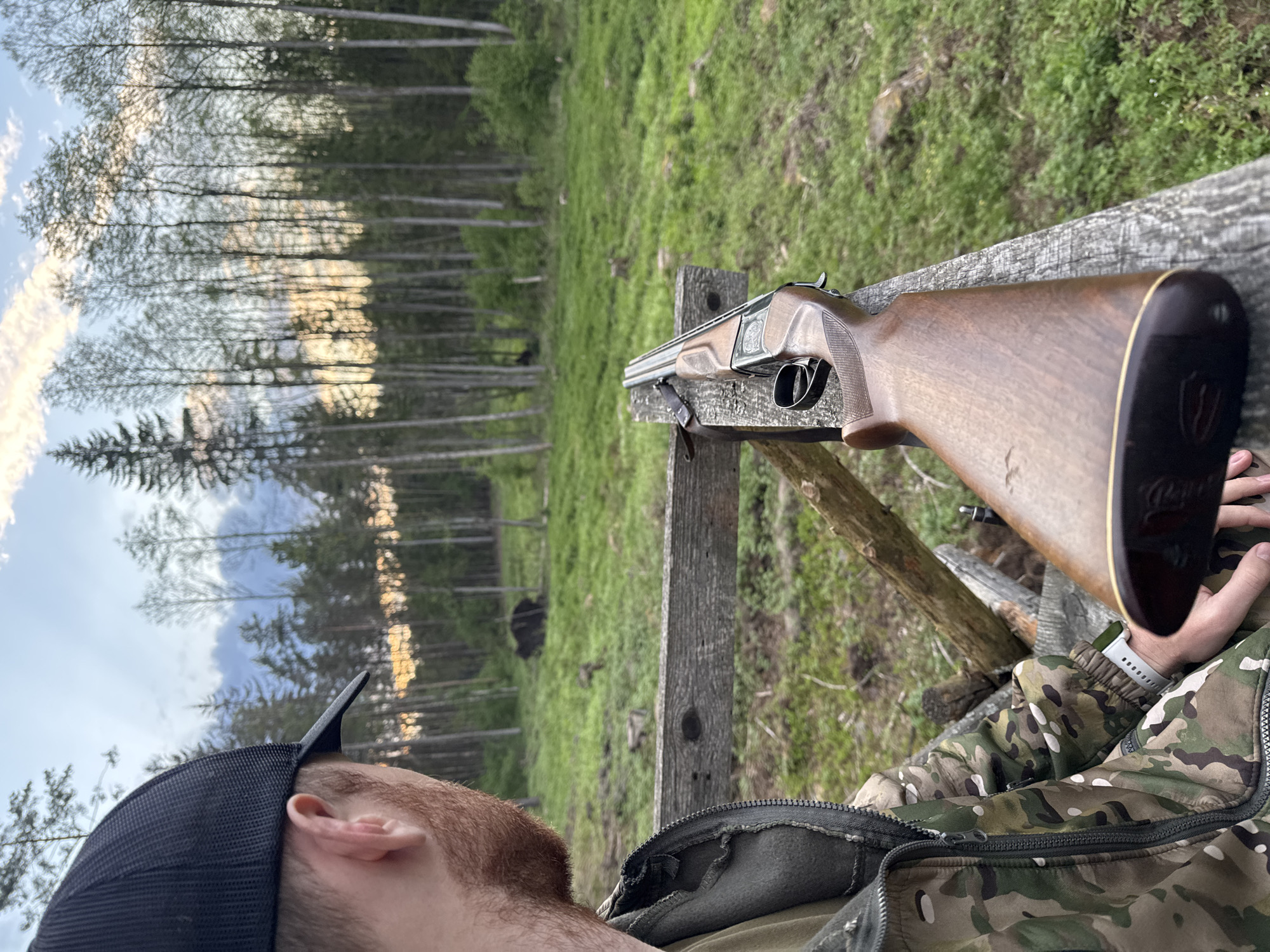 The image shows a man wearing a camouflage jacket and a black baseball cap, with a partial view of his face and beard. A wooden double-barreled shotgun rests on a weathered wooden railing. The background consists of a grassy field and a forest of thin trees stretching towards a sky with clouds and a hint of sunset. The location appears to be a rural or wooded area, but there is no specific indication of a city.