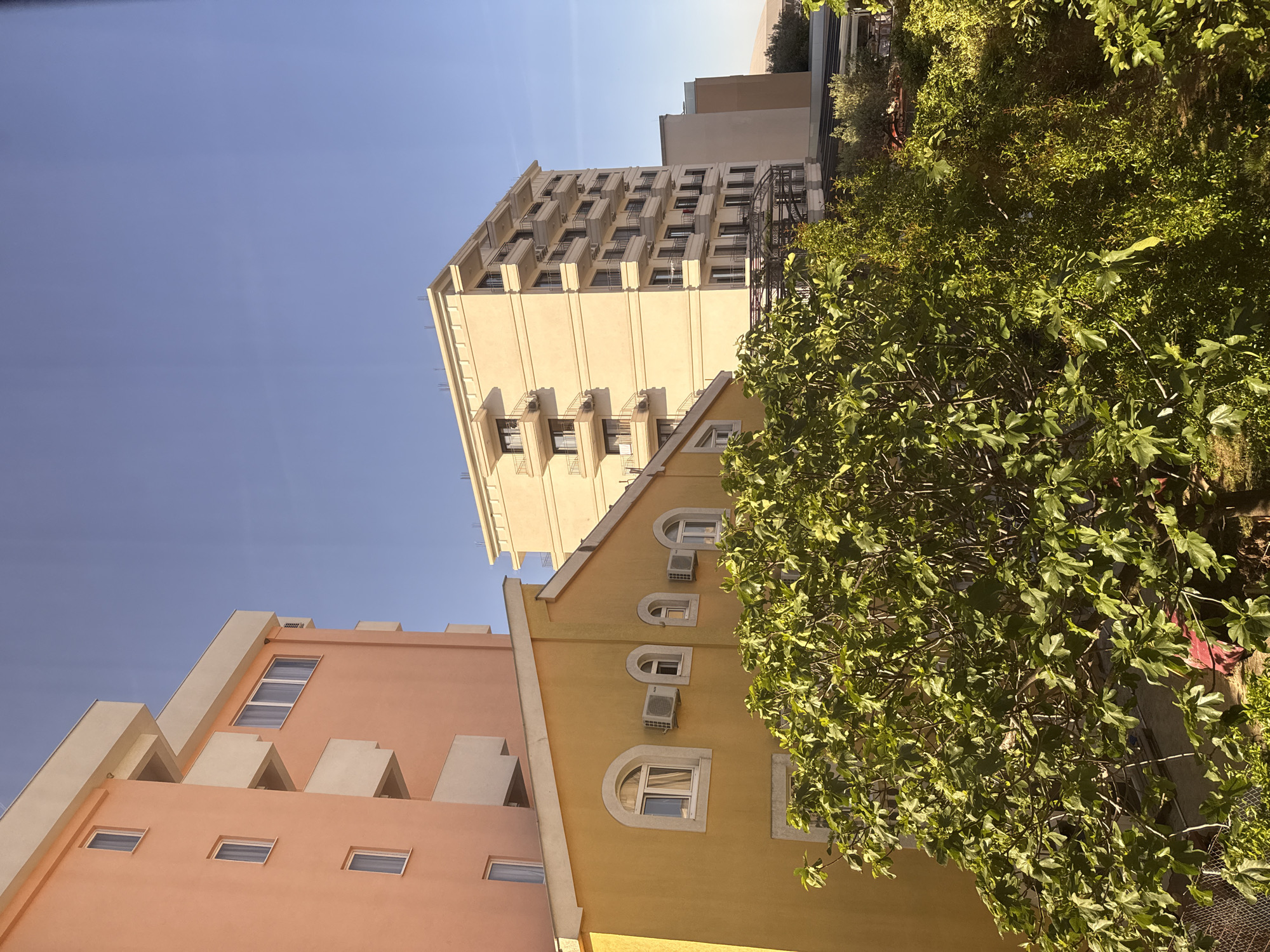 The image shows a view of buildings in Bar, Montenegro. A peach-colored building is on the left, a yellow building in the center with arched windows, some of which have air conditioning units. Behind those, a taller beige building rises several stories with balconies. Part of a tree or bush is visible on the right. The sky is blue and clear.