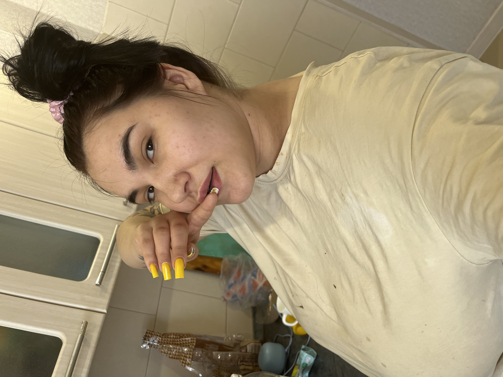 A woman with dark hair pulled into a high bun with a pink scrunchie is posing indoors. She is wearing a light-colored t-shirt with visible stains. Her fingernails are long and painted a bright yellow. She has a tattoo on her hand. The background shows a kitchen area with cabinets and various items on the counter. The location is Chaikovsky, Russia, based on the available information.
